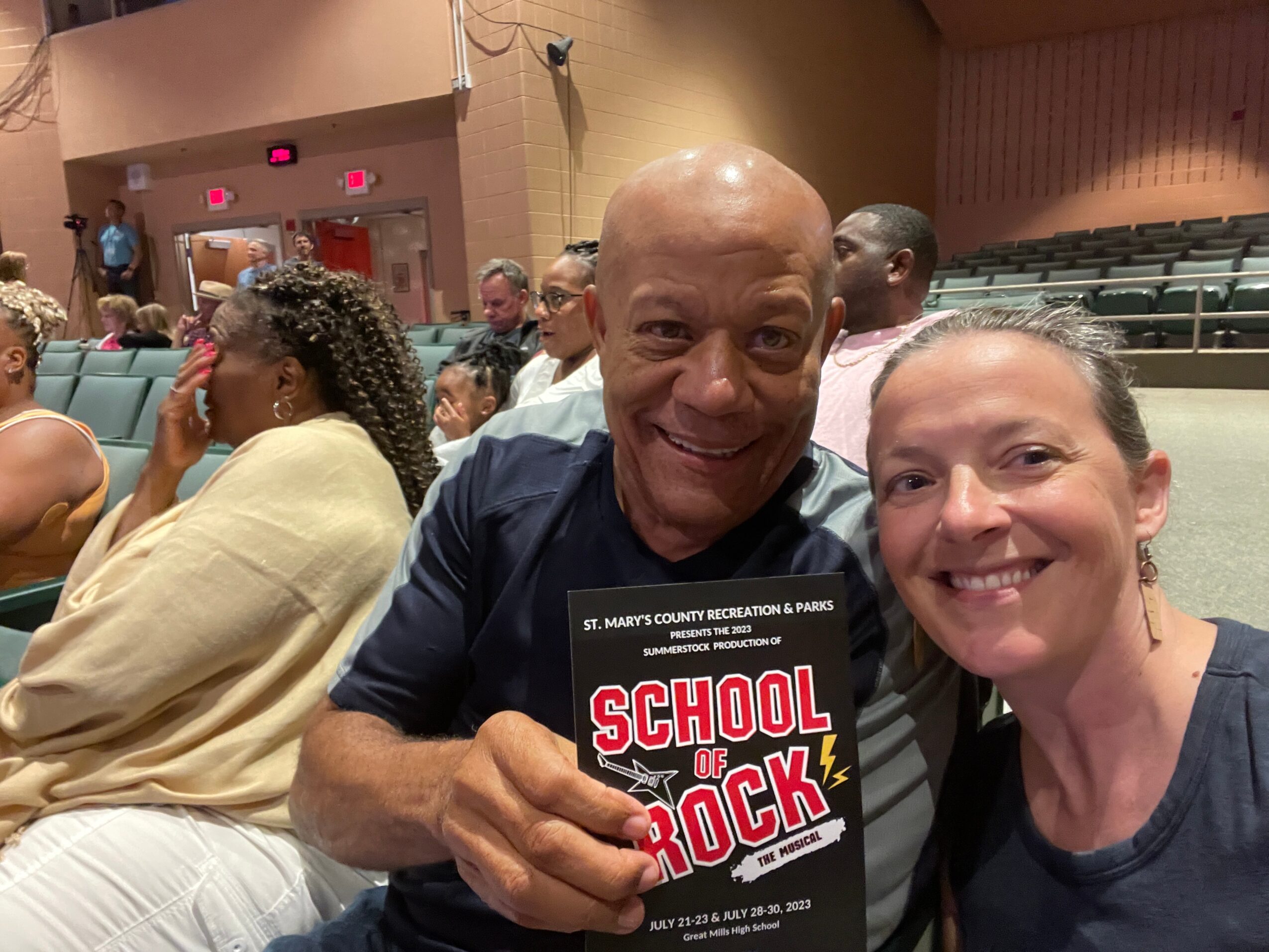 Two people smiling, one holding a sign that says 'School of Rock,' in an indoor auditorium with other attendees.