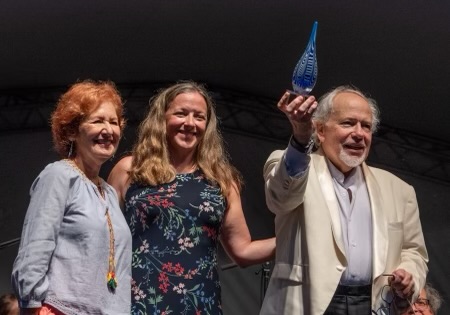 Three people on stage, one holding a blue glass award, smiling, with a dark background.