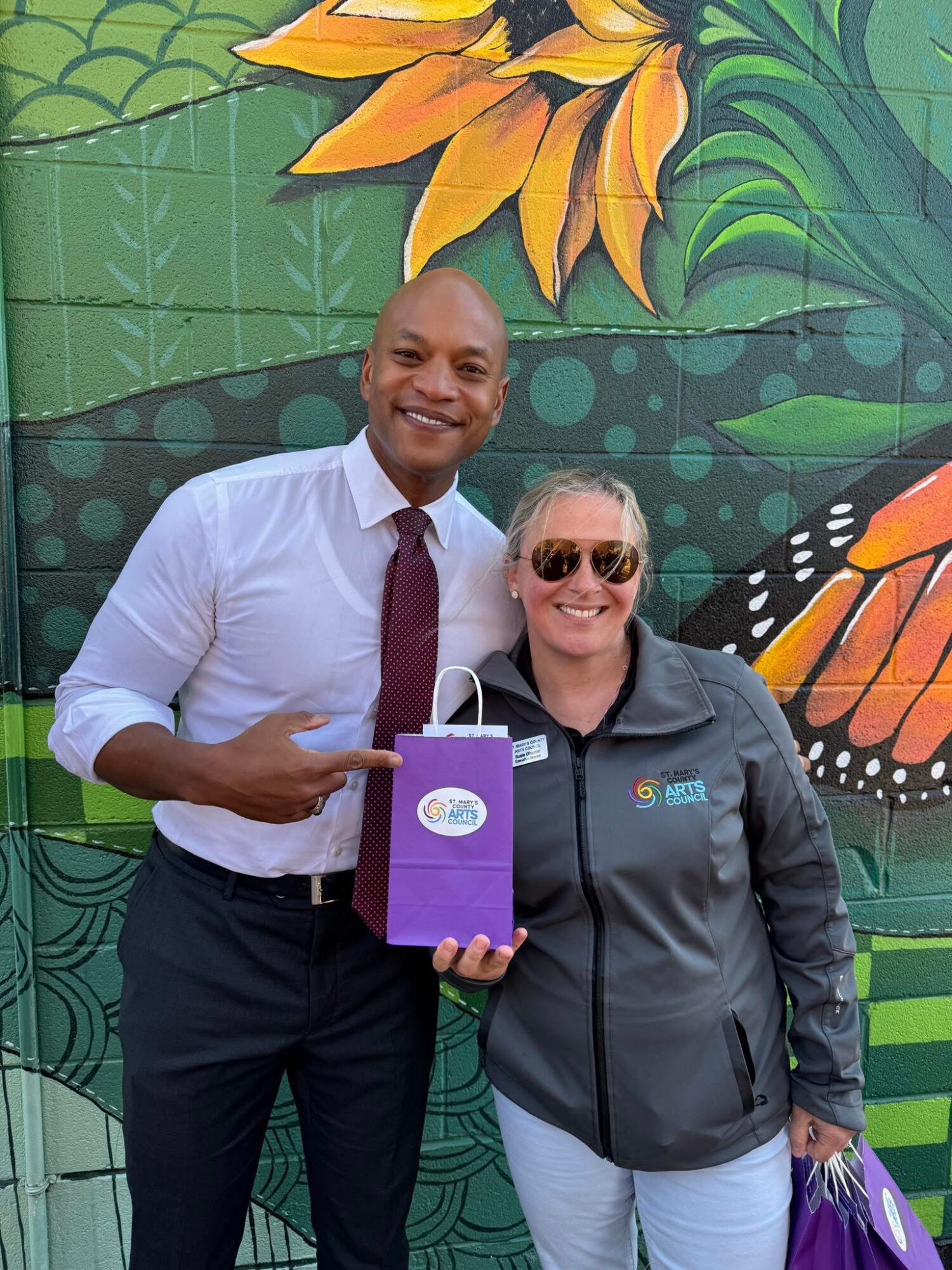Two people smiling, holding a purple gift bag, standing in front of a colorful mural with a sunflower and butterfly design.