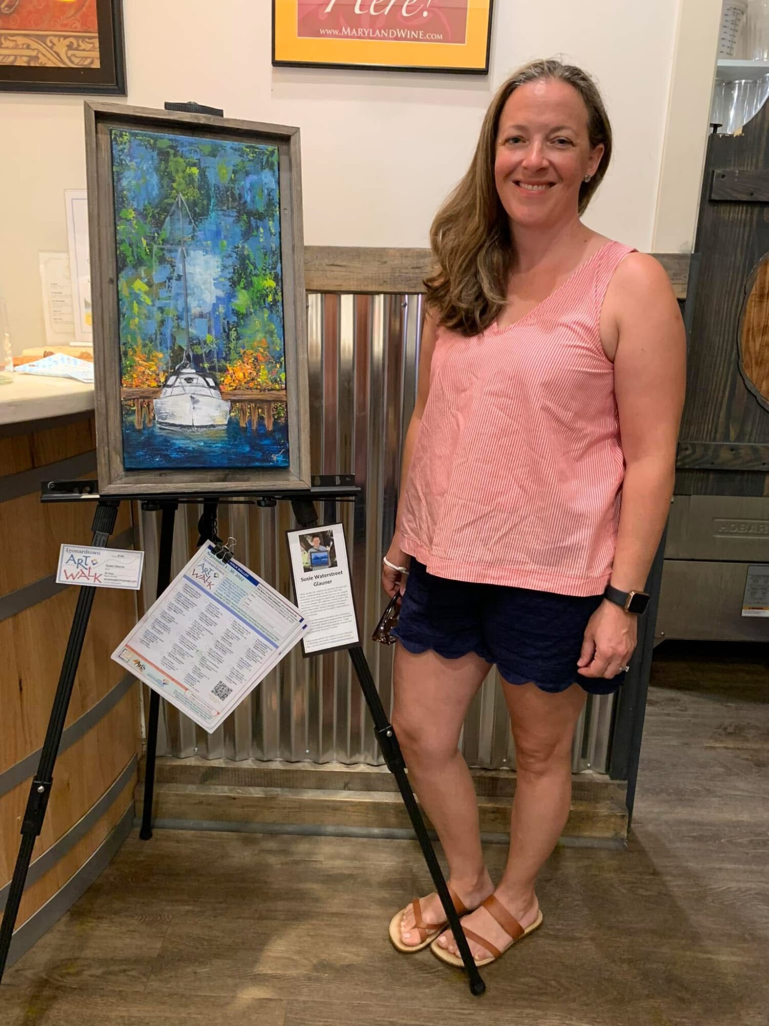 Woman in pink top and dark shorts standing next to a colorful painting on an easel in an indoor setting.