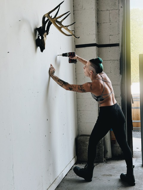 Person with tattoos and green hair pushes a mounted deer head on a white wall in a room with concrete walls and a window.