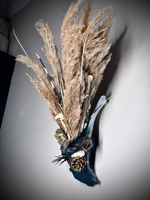 Decorative wall piece with dried pampas grass, a pine cone, and a curved blue element, mounted on a gray wall.