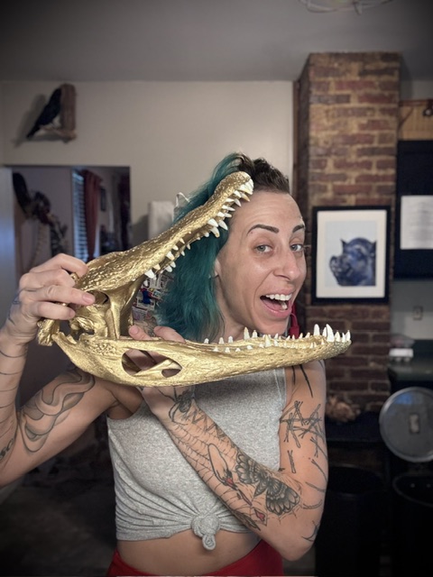 Woman with teal hair holding a crocodile skull prop, smiling in a room with brick wall and framed pictures.