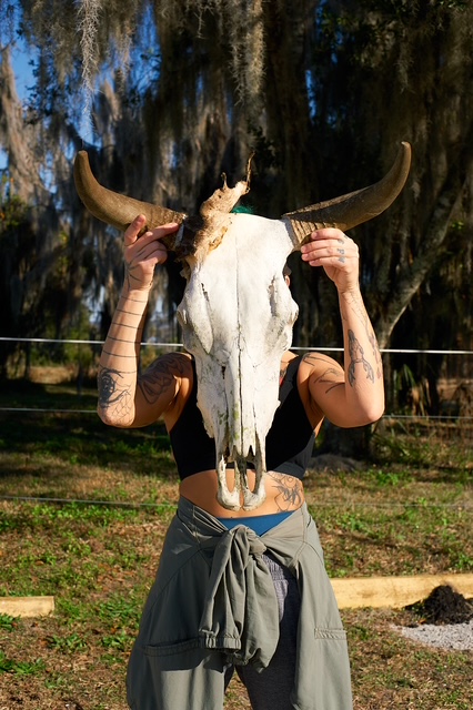 Person holding a large animal skull with horns in front of their face outdoors, trees and grass in background.