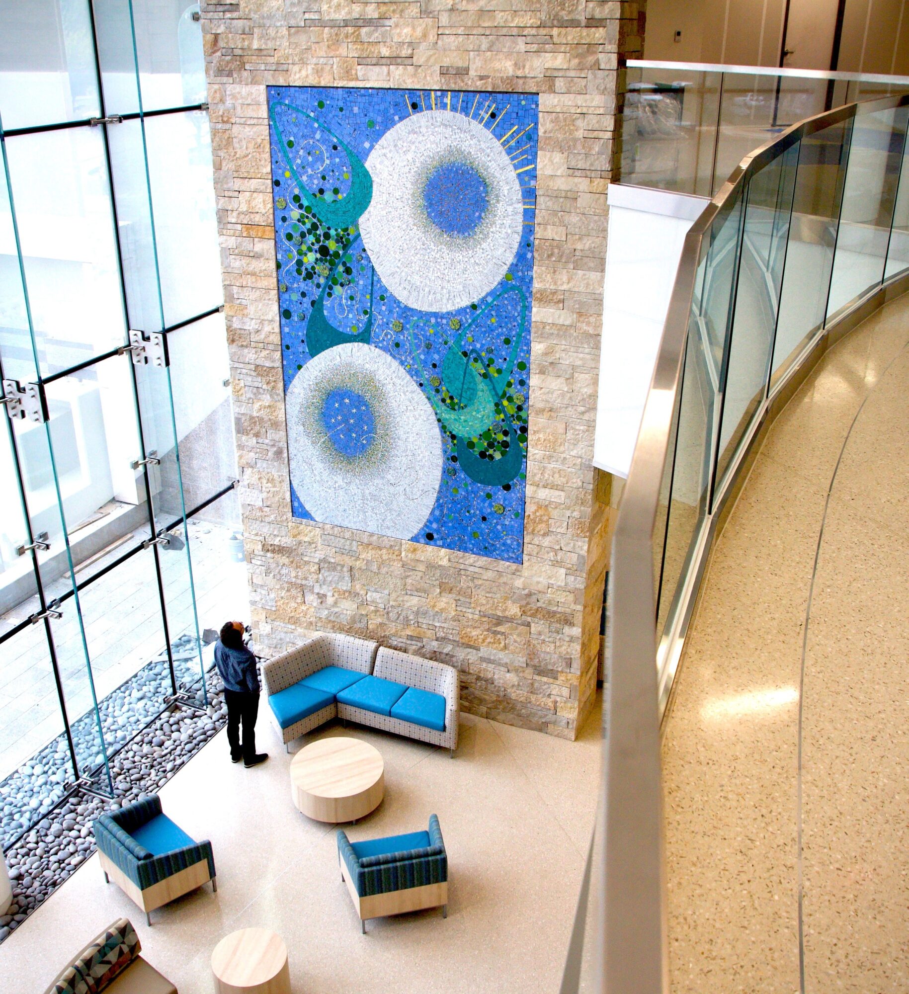 Interior view of a modern building with a large abstract artwork on a brick wall, seating area, and a person standing nearby.