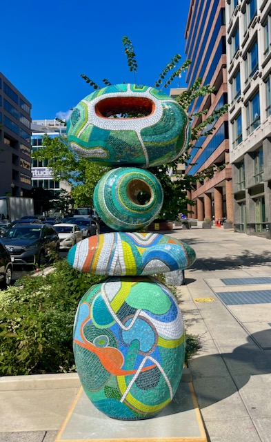 Colorful mosaic sculpture of stacked abstract shapes on a city sidewalk with buildings and cars nearby.