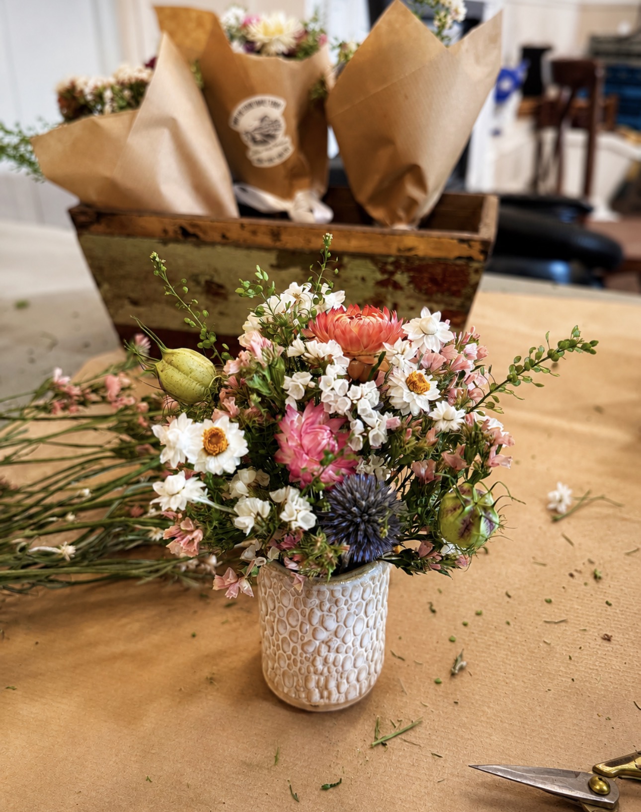Colorful mixed flower bouquet in a textured white vase on a wooden table, with a wooden box of wrapped flowers in the background.