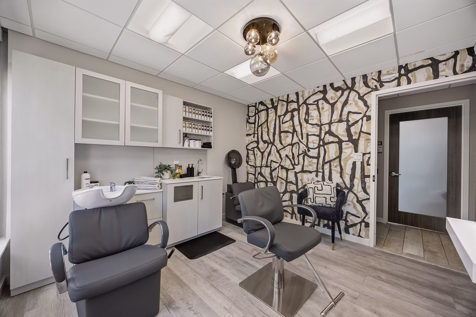 Office room with white cabinets, a patterned wall, two gray chairs, and a door, with a ceiling light fixture.