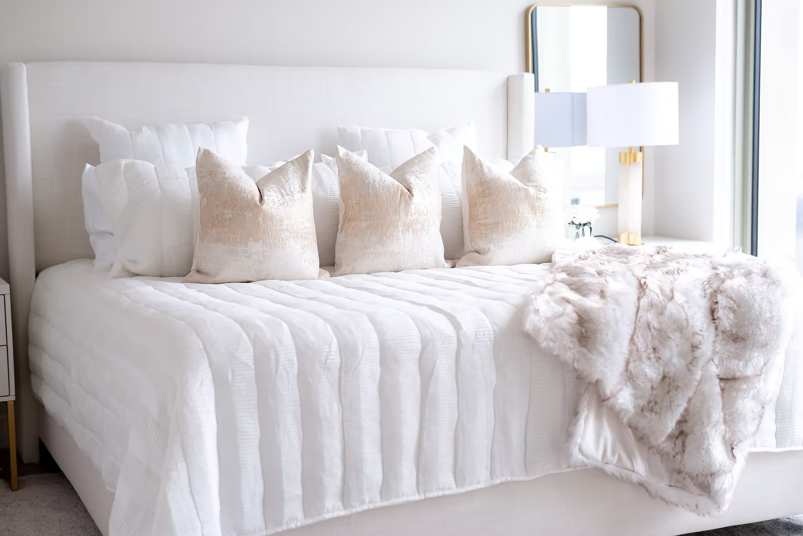 Upright bed with white bedding, multiple pillows, and a plush throw blanket, next to a window with a lamp and mirror.