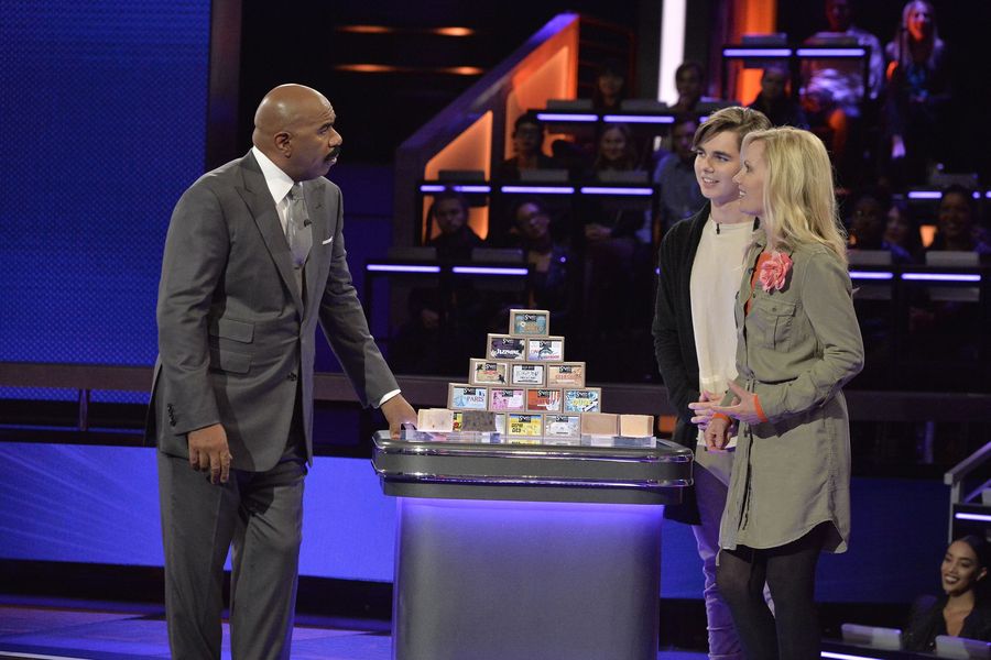 Three people stand around a table with stacked boxes on a stage, with an audience in the background.