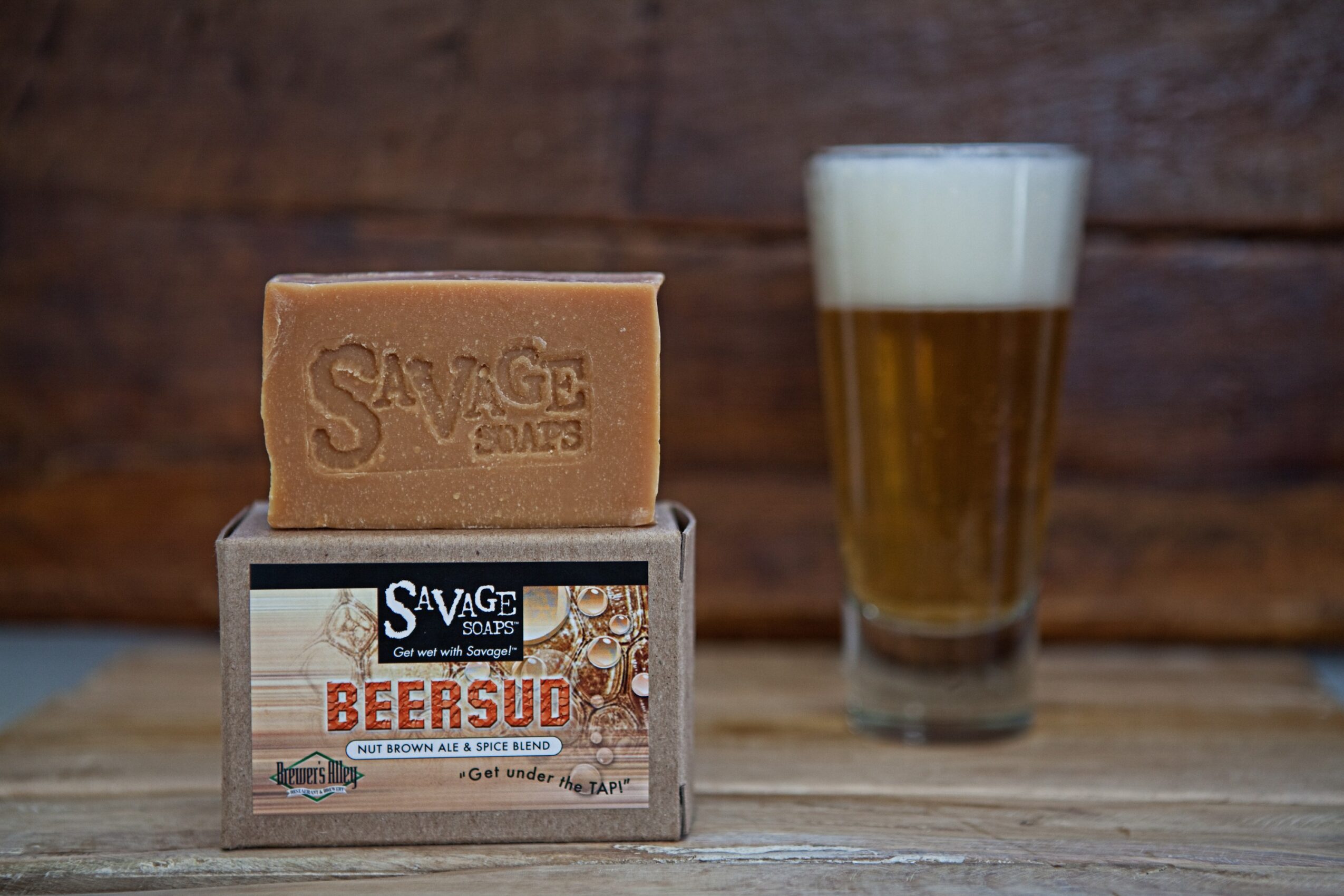 Bar of soap labeled 'Savage' on a small box, with a glass of beer in the background, on a wooden surface.