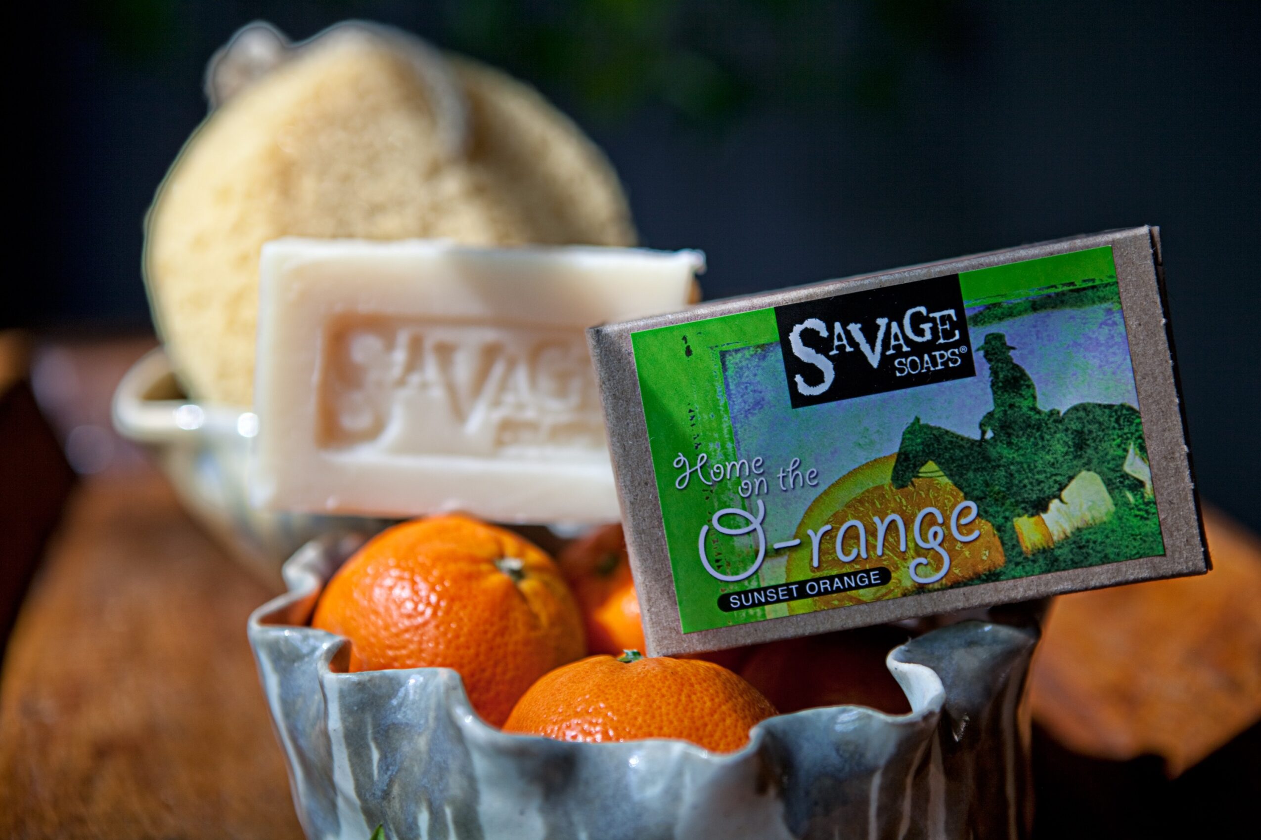Bar of Savage soap, oranges, and a loofah on a wooden surface, with a soap dish in the background.