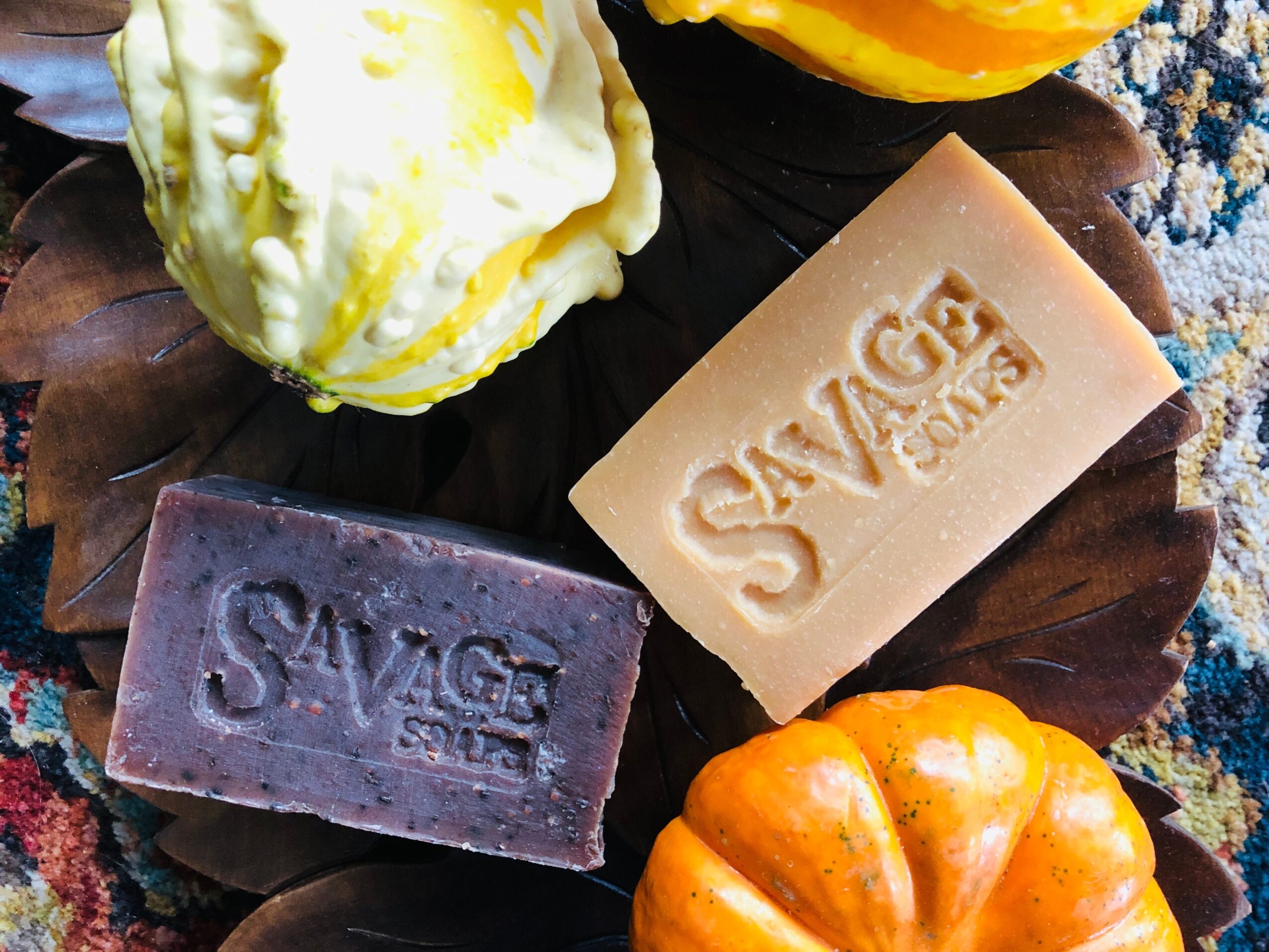 Three bars of soap with 'SAVAGE' embossed, one yellow and one purple, on a wooden surface with a yellow flower nearby.