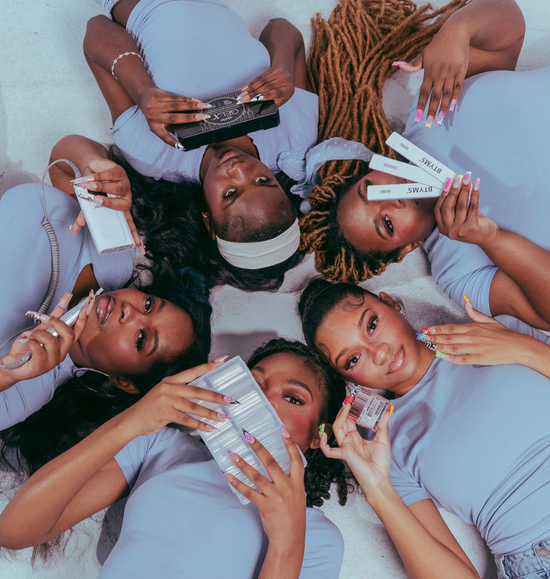 Six women lying on their backs in a circle, holding various beauty products, wearing light blue outfits, looking at the camera.