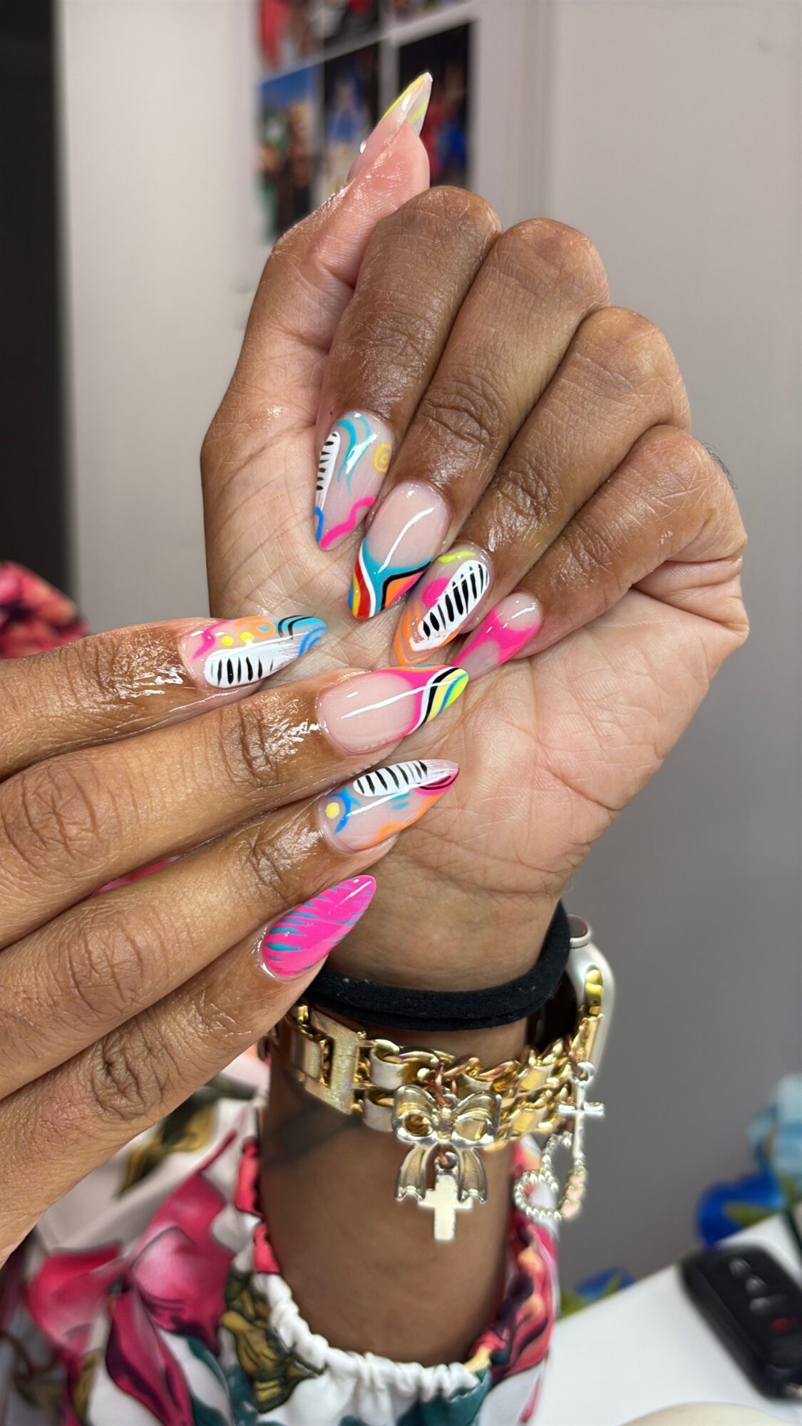 Person's hand with colorful, striped nail art, wearing multiple bracelets, including a gold chain and charms, against a blurred background.