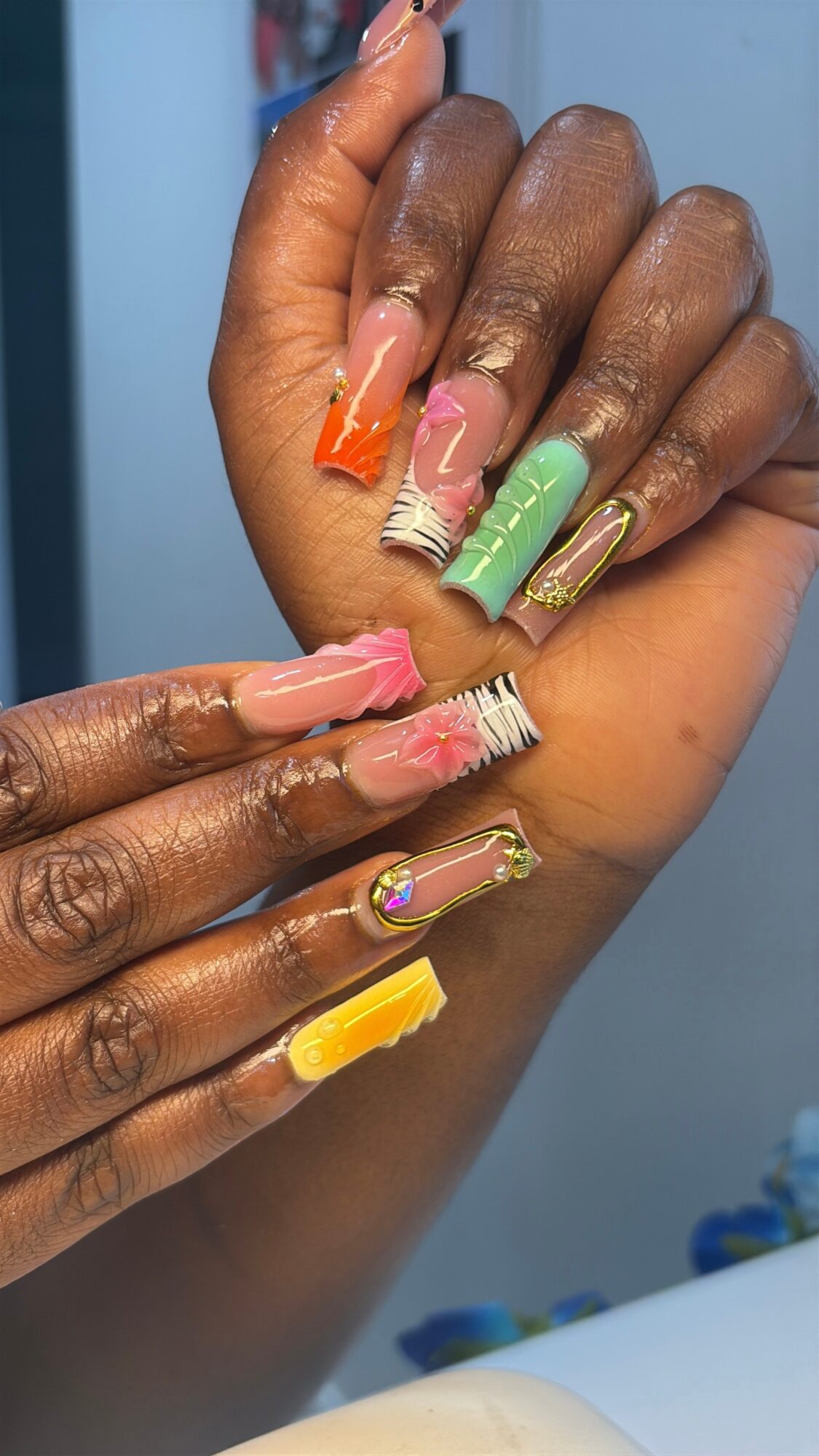 Two hands with colorful, decorated nails, some with floral and geometric designs, against a blue background.