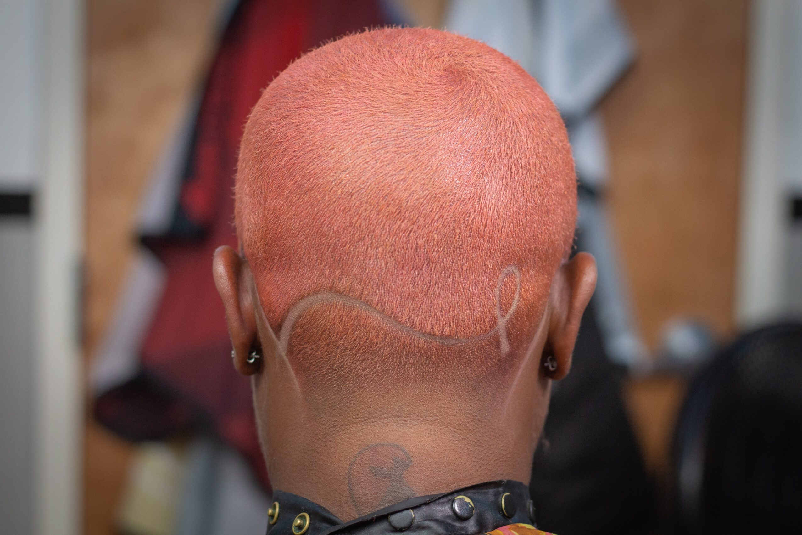 Back of a person's head with short, pink-colored hair, earrings, and a tattoo on the neck, in a room with clothing and objects.