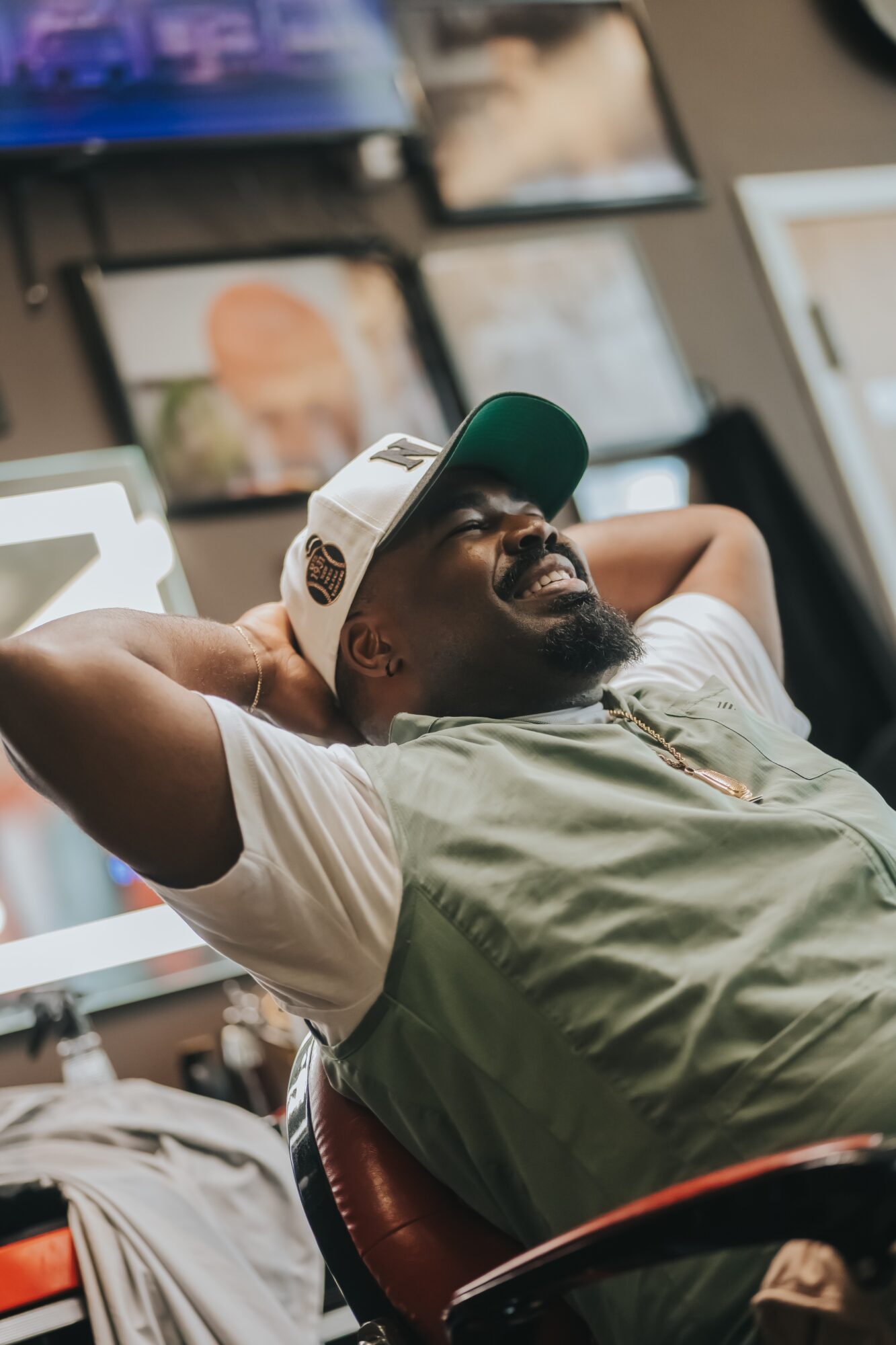 Man with a beard and glasses, wearing a cap, relaxing with arms behind head, smiling, surrounded by screens and devices.