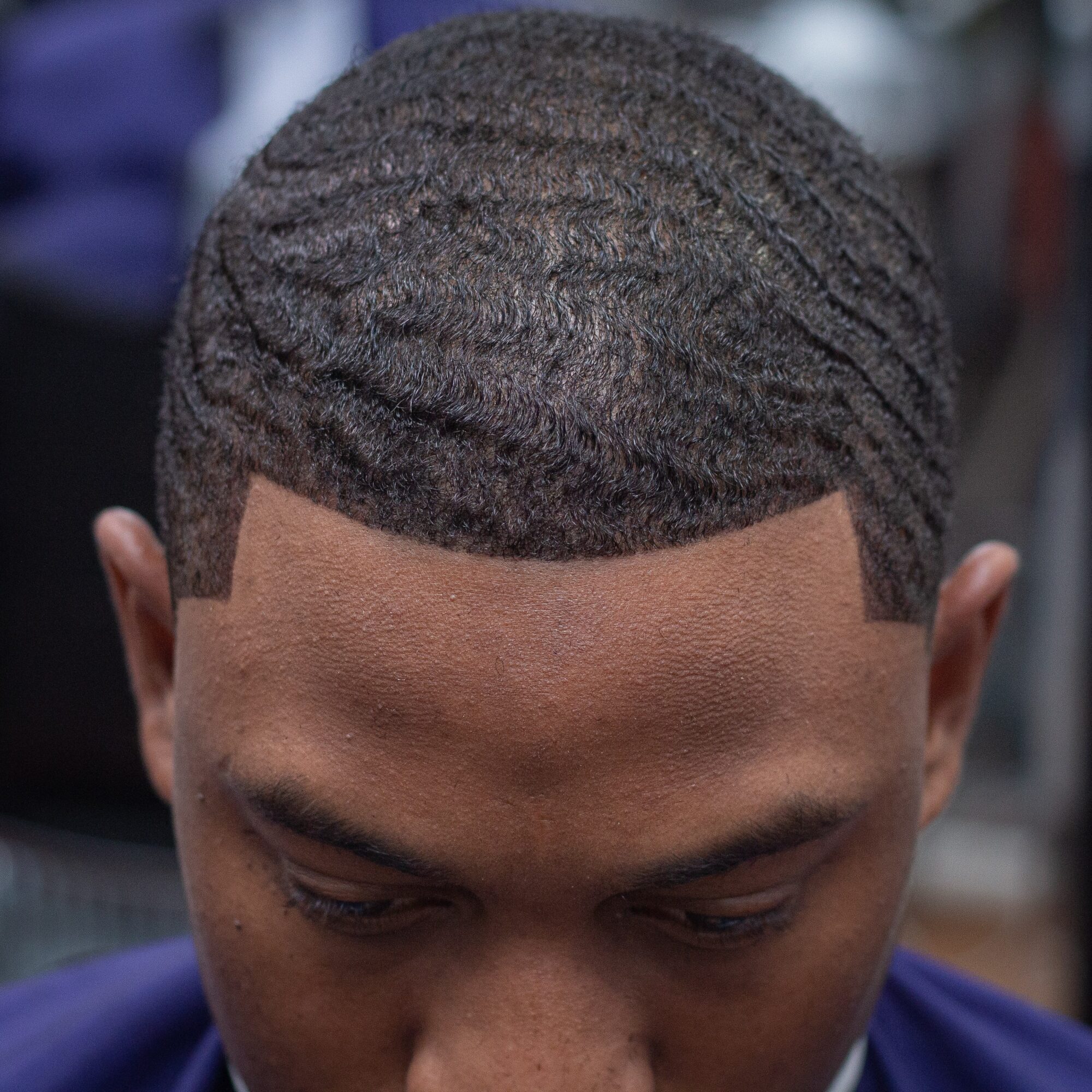 Close-up of a person's head with short, wavy hair and a defined hairline, looking downward.