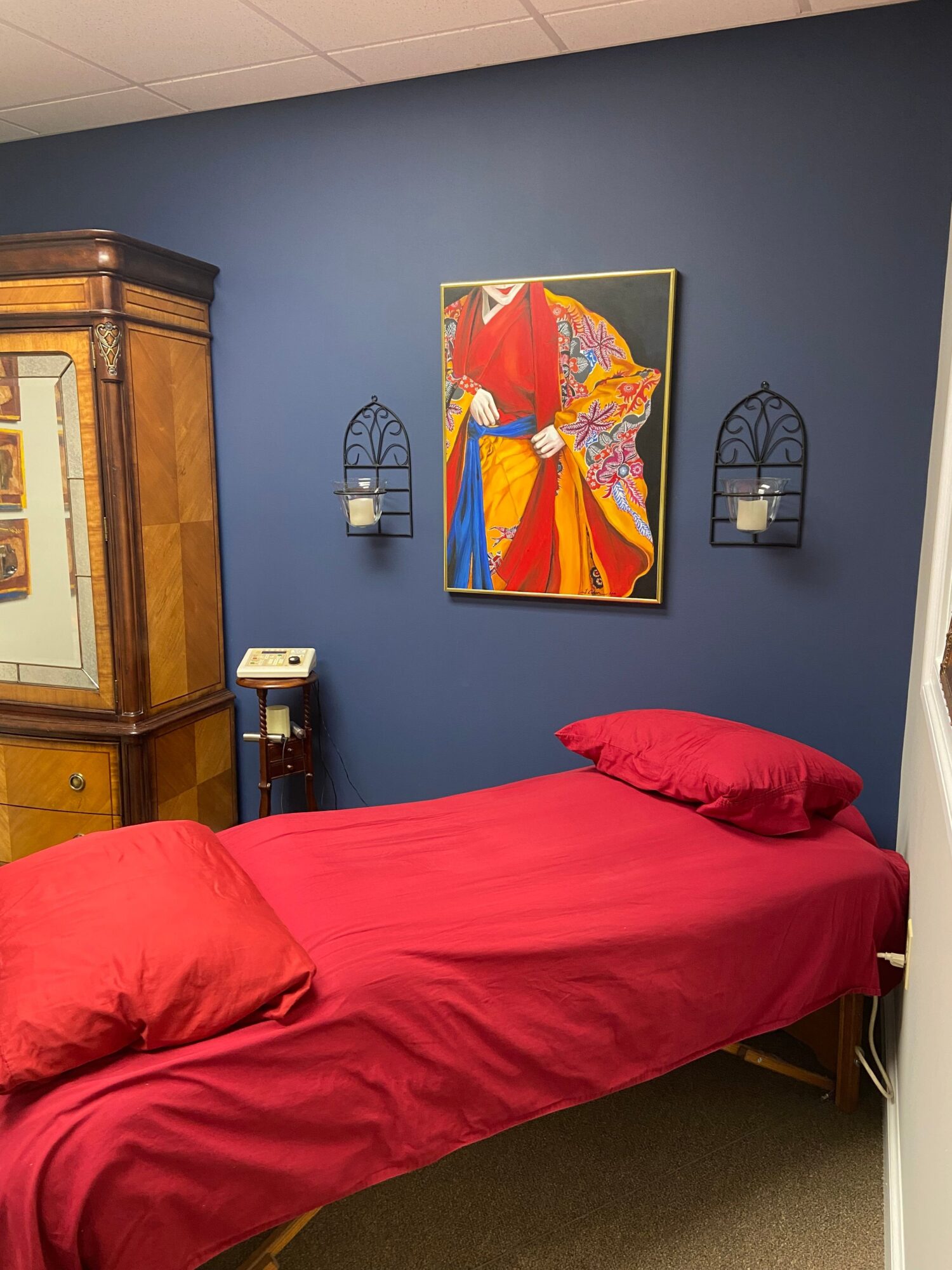 A bedroom with a blue wall, a colorful mountain painting, a red bed, and a nightstand with a lamp and clock.