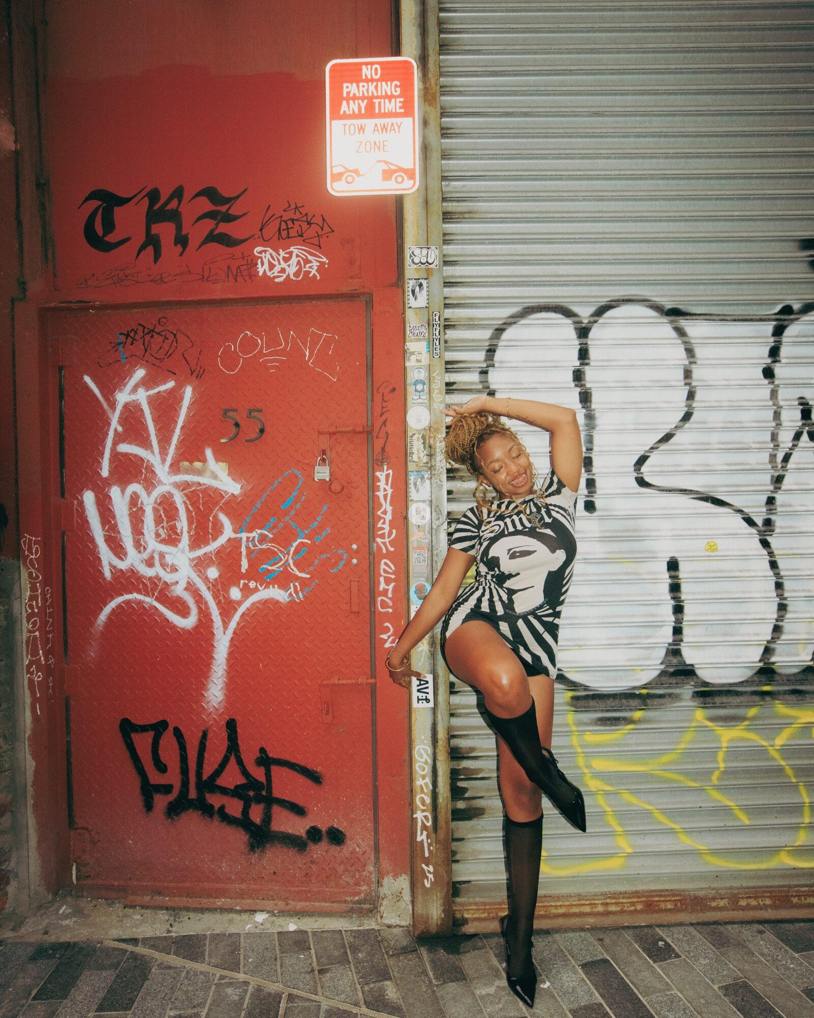Person with curly hair poses against a red door and graffiti-covered wall, wearing a striped top, shorts, and high boots.