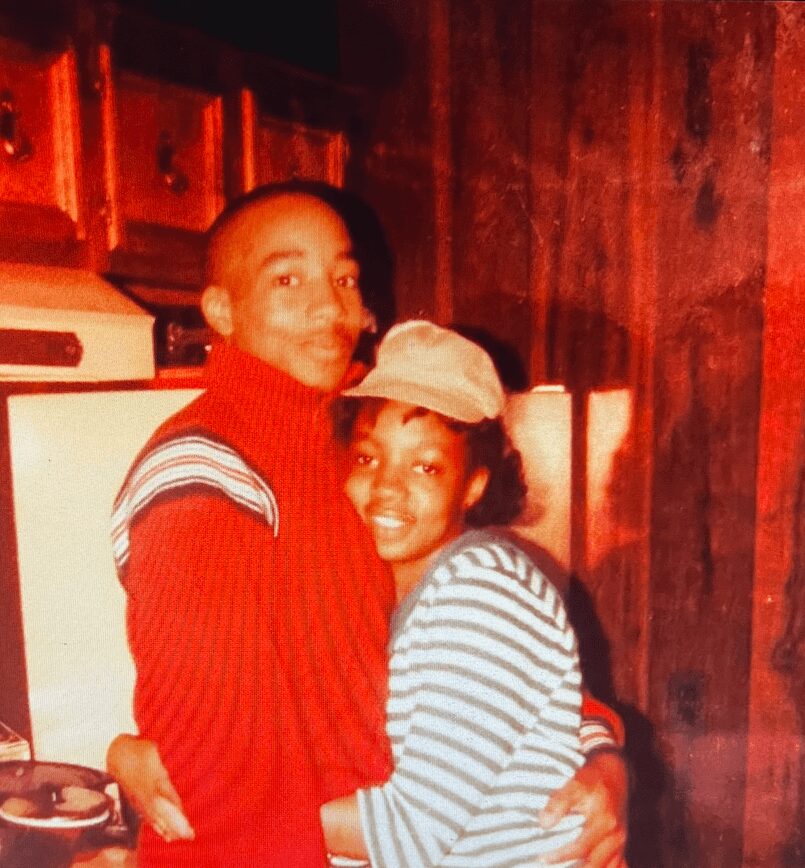 Two people hugging indoors, one wearing a red sweater and the other a striped shirt and cap, wooden wall background.