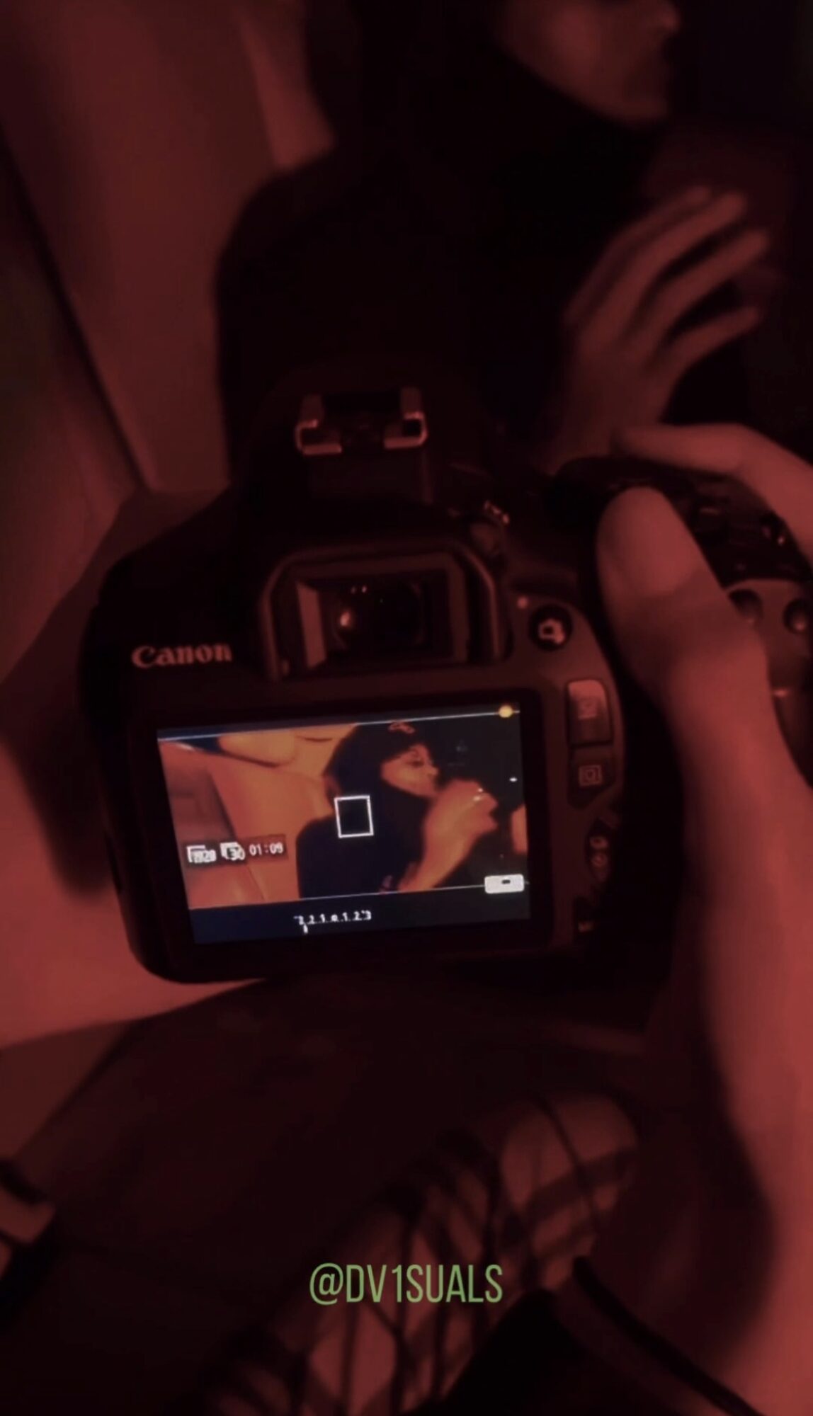 Person holding a camera, reflection of person in camera screen, dark background, red lighting, hand near face, other person in background