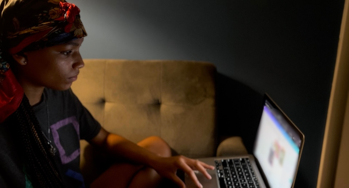 Person with headscarf using a laptop on a sofa in a dimly lit room.
