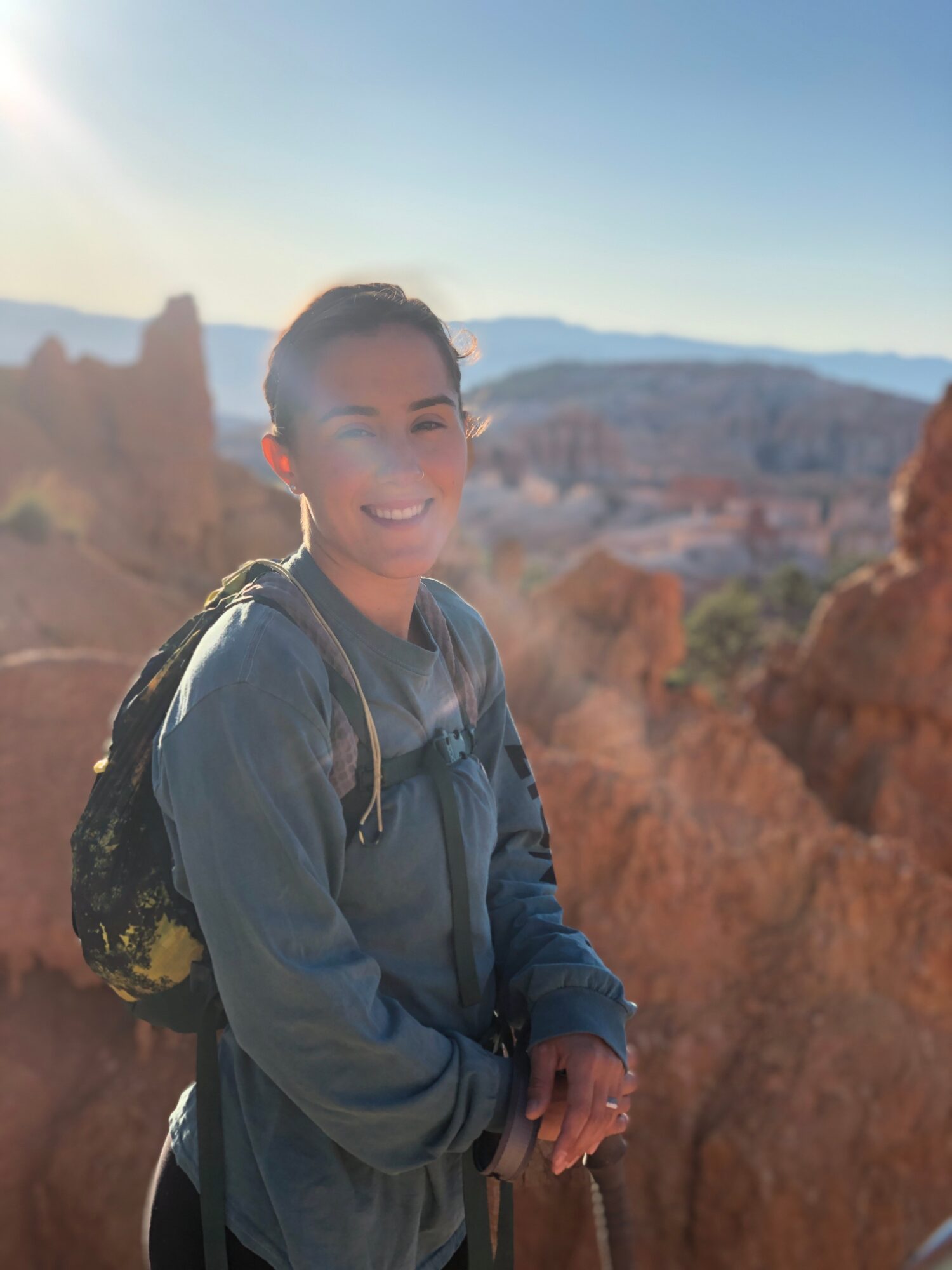 Smiling woman with backpack outdoors in a rocky landscape, sunlight shining, blue sky, distant mountains.