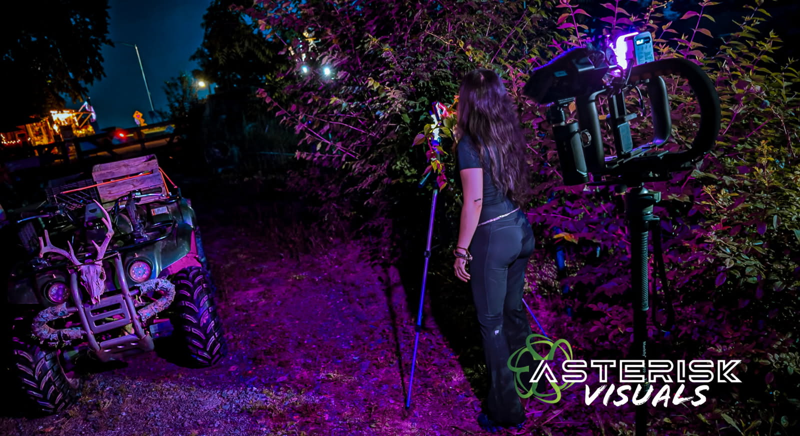 Person with long hair holding a stick stands outdoors at night near a vehicle and bushes, illuminated by purple light.