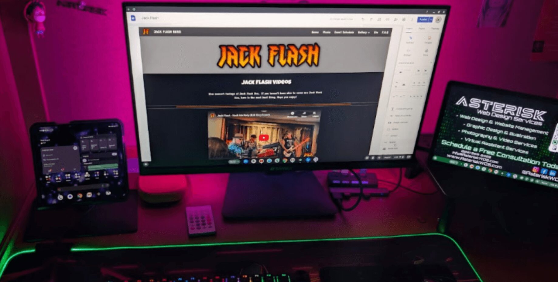 Computer monitor displaying 'Jack Flash' website, tablet showing 'Asterisk' game, keyboard with green backlight, and smartphone on desk.