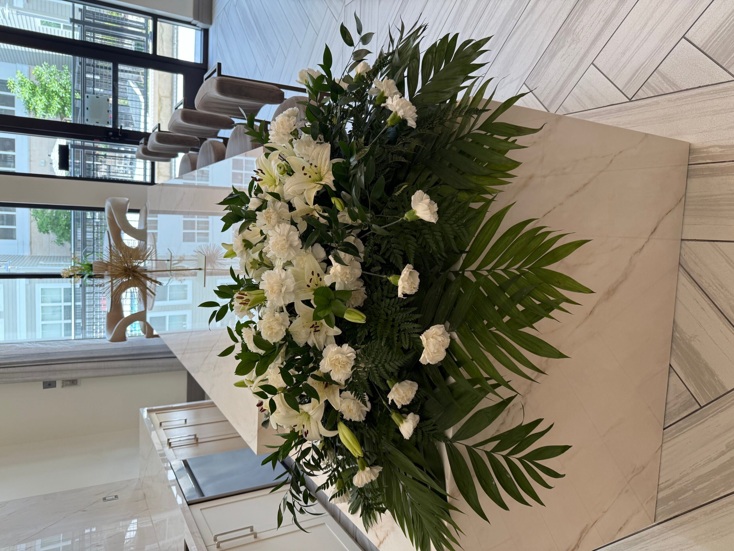 Large floral arrangement with white flowers and green leaves on a marble table in a bright room.