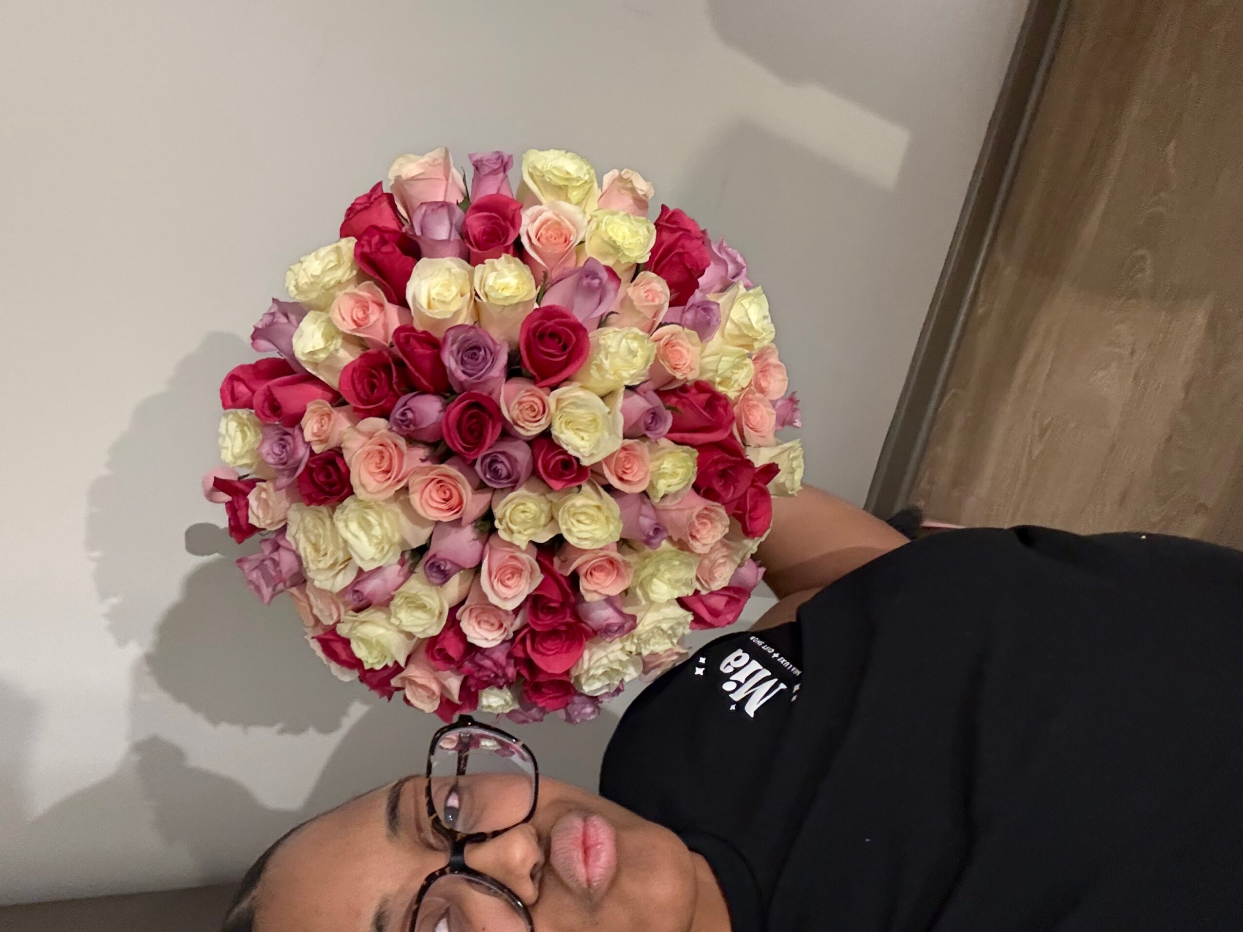 Person holding a large bouquet of pink, white, and red roses, wearing glasses and a black shirt, standing indoors.