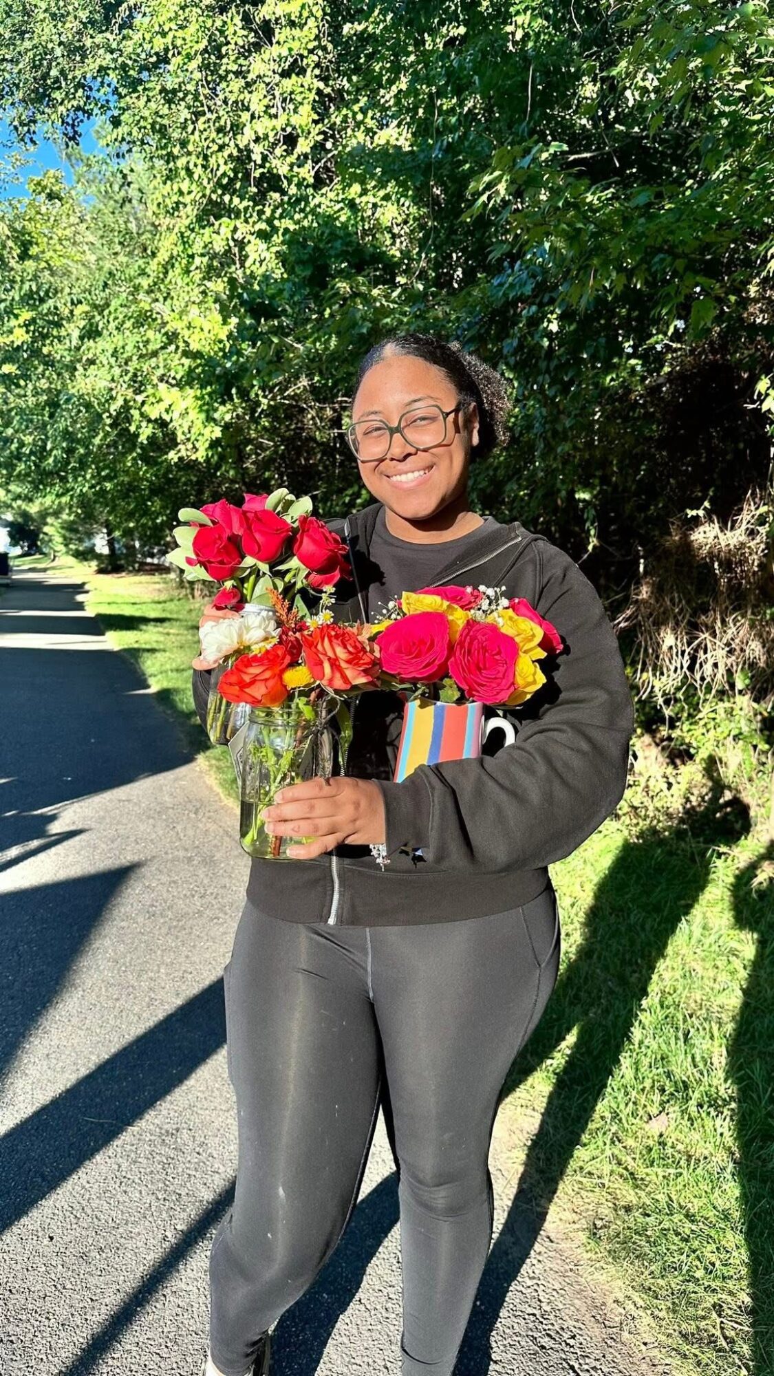 Woman holding a bouquet of red and pink flowers outdoors, smiling, with trees and a sidewalk in the background.