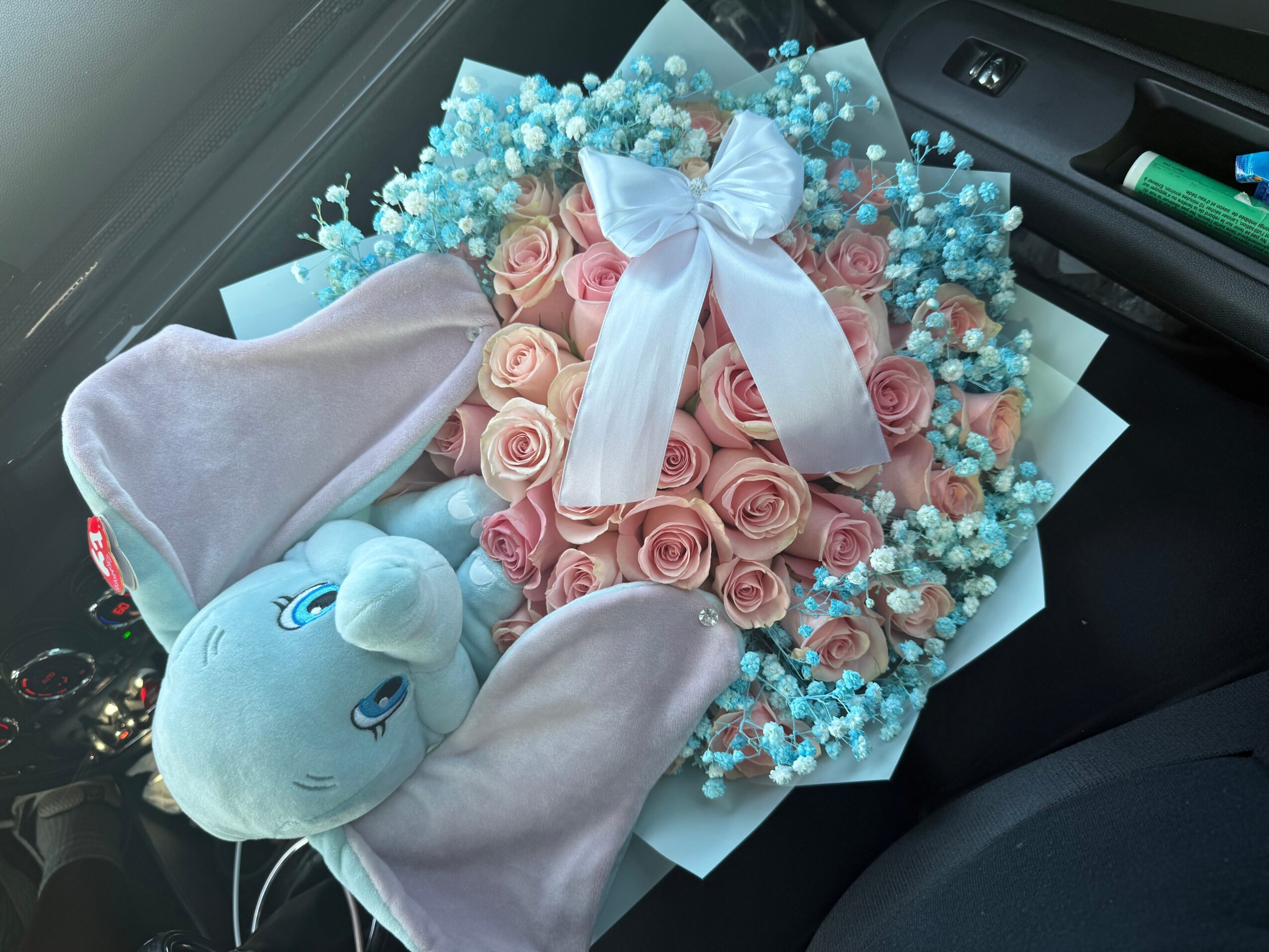 A bouquet of pink roses, blue baby's breath, a white ribbon, and a plush bunny toy inside a car. Alt text word count: 20