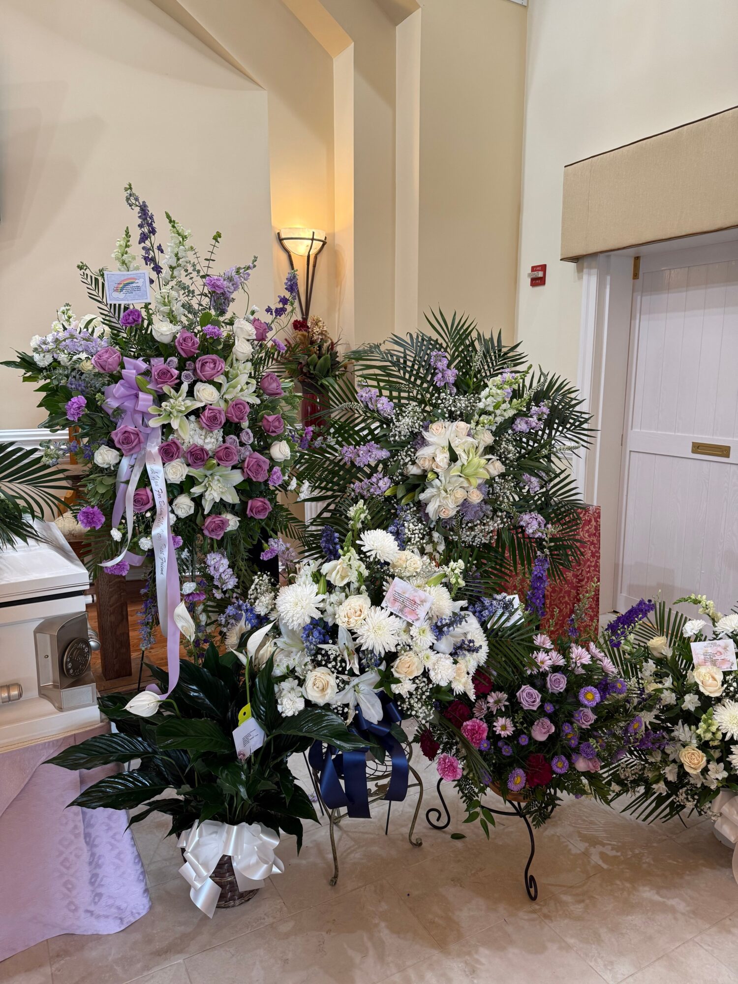 Multiple floral arrangements with white, purple, and pink flowers on a table, with a wooden floor and white wall in background.