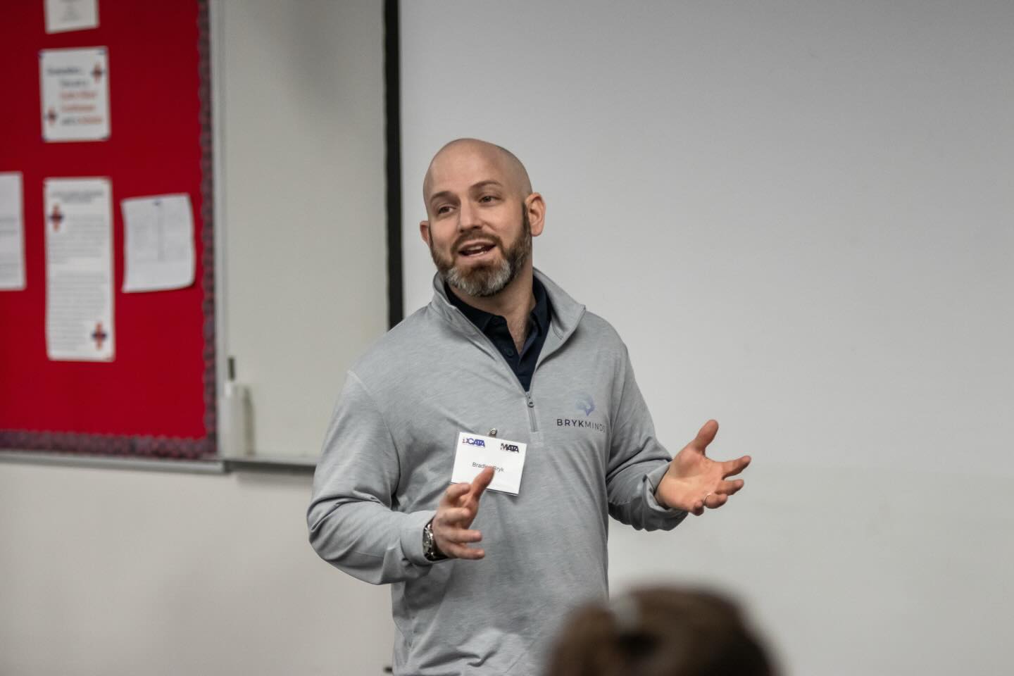 Man with a beard and bald head speaking and holding a card in a room with a white wall and red bulletin board