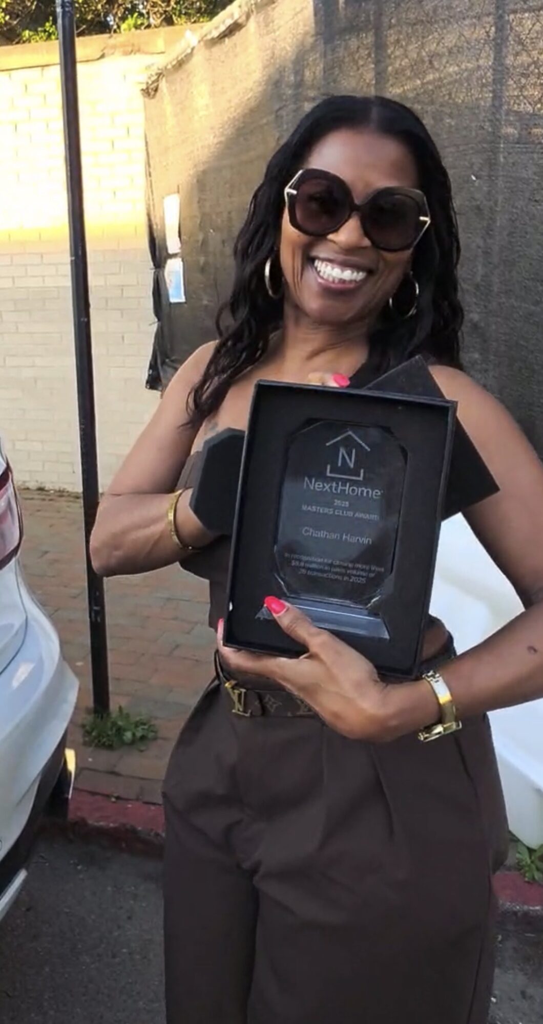 Woman with sunglasses holding an award plaque outdoors, smiling, wearing a black top and pants, with a brick wall background.