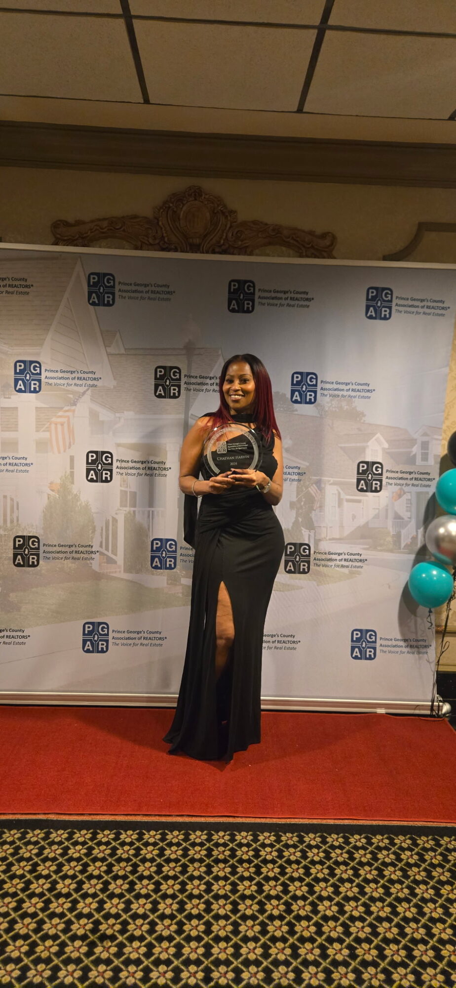 Woman in a long dress holding an award, standing on a red carpet in front of a backdrop with logos.