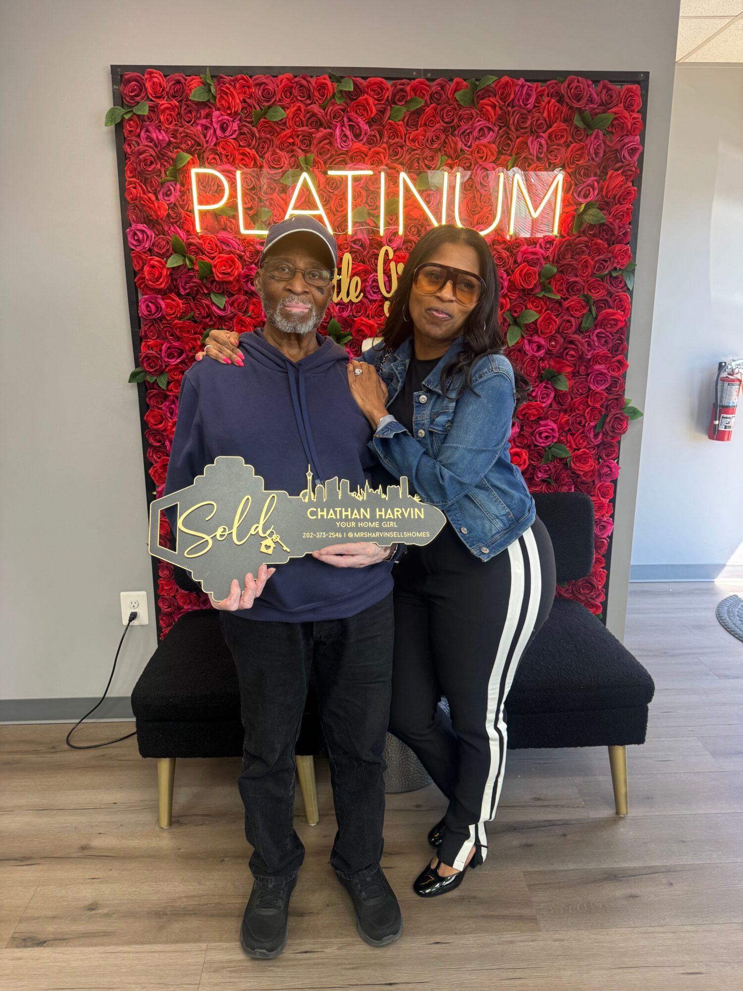Two women standing in front of a red floral backdrop, smiling, one holding a plaque, in a room with wooden floor.