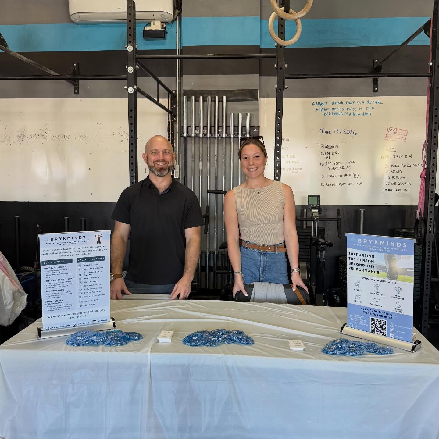 Two people standing behind a table with informational flyers, in a gym setting with rings and a whiteboard in the background.
