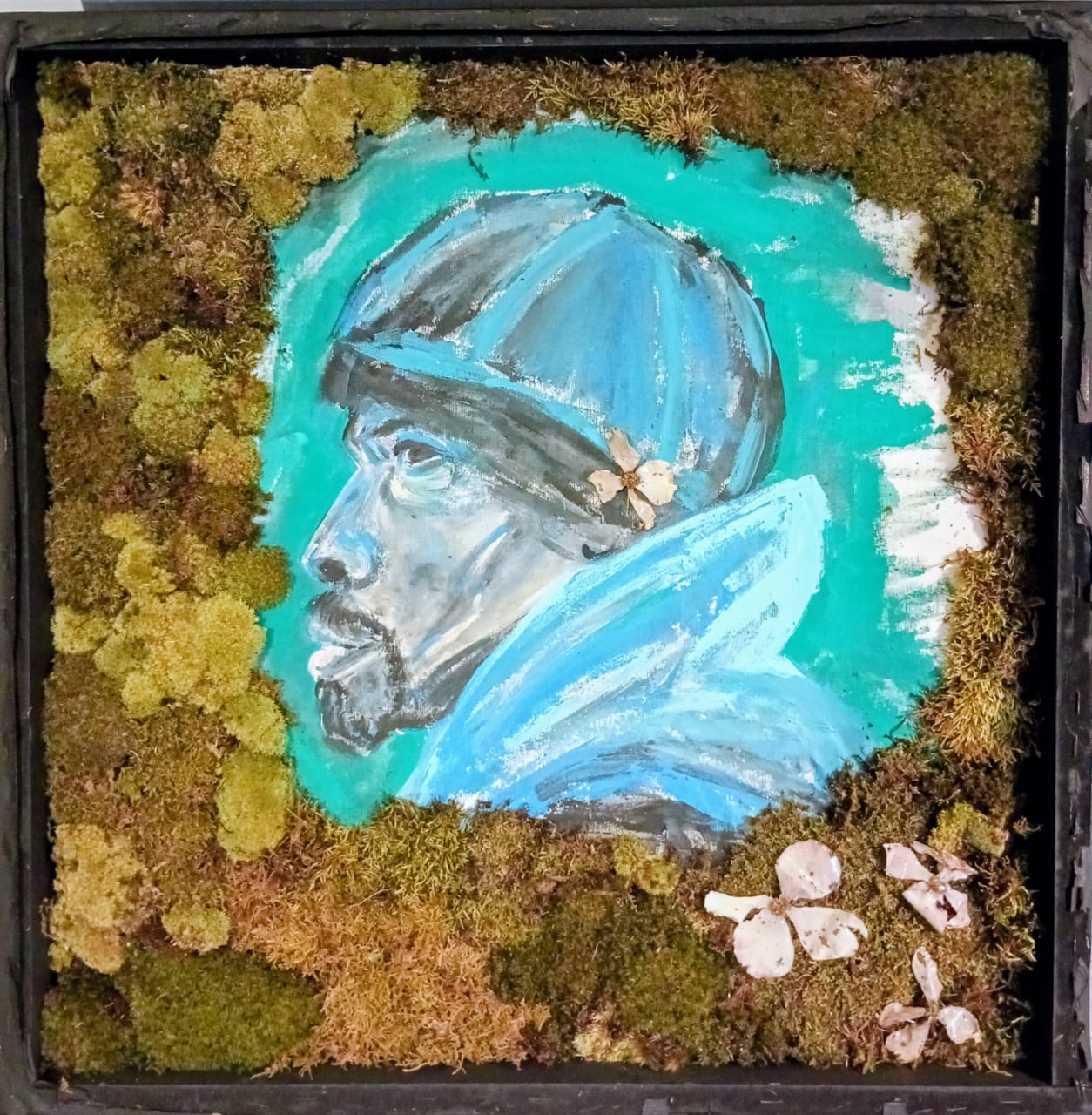 Painting of a person's profile with a blue hat, surrounded by moss and small white flowers, in a black frame.