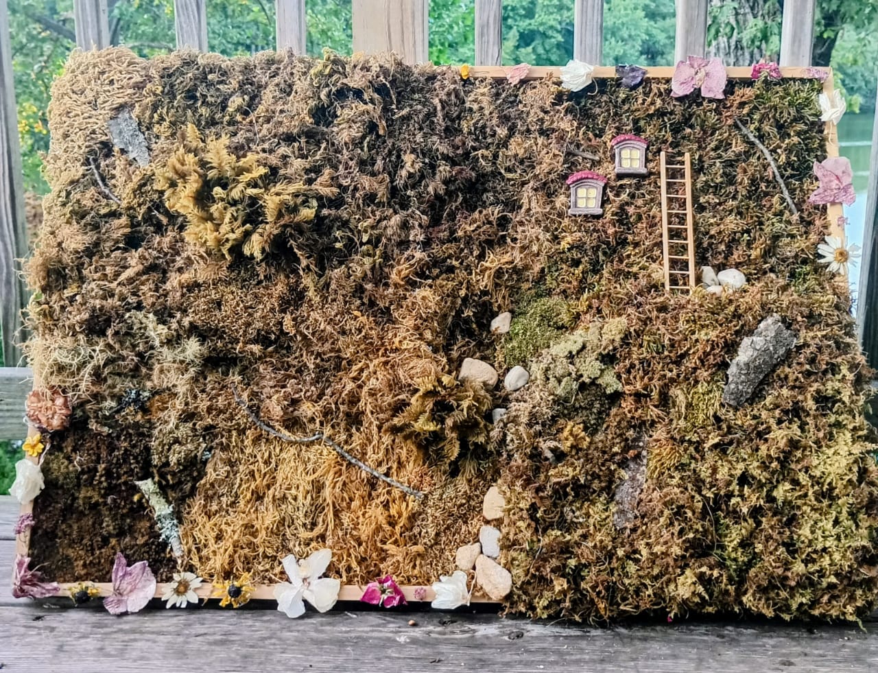 A textured mossy landscape with a ladder, small structures, and flowers, resembling a miniature scene or diorama.