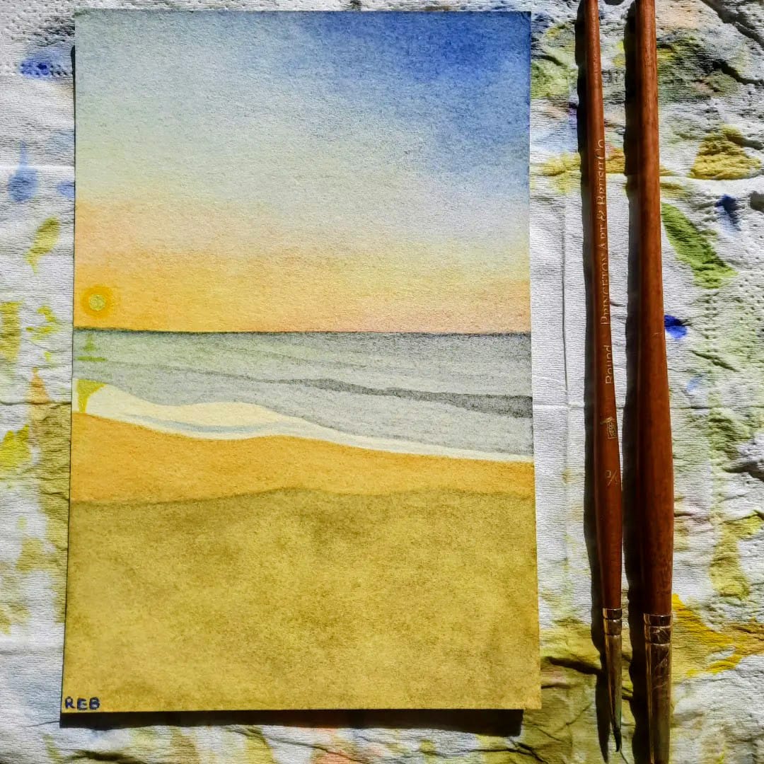 Watercolor painting of a beach scene with sky, ocean, and sandy shore, placed on a textured surface with two wooden brushes beside it.