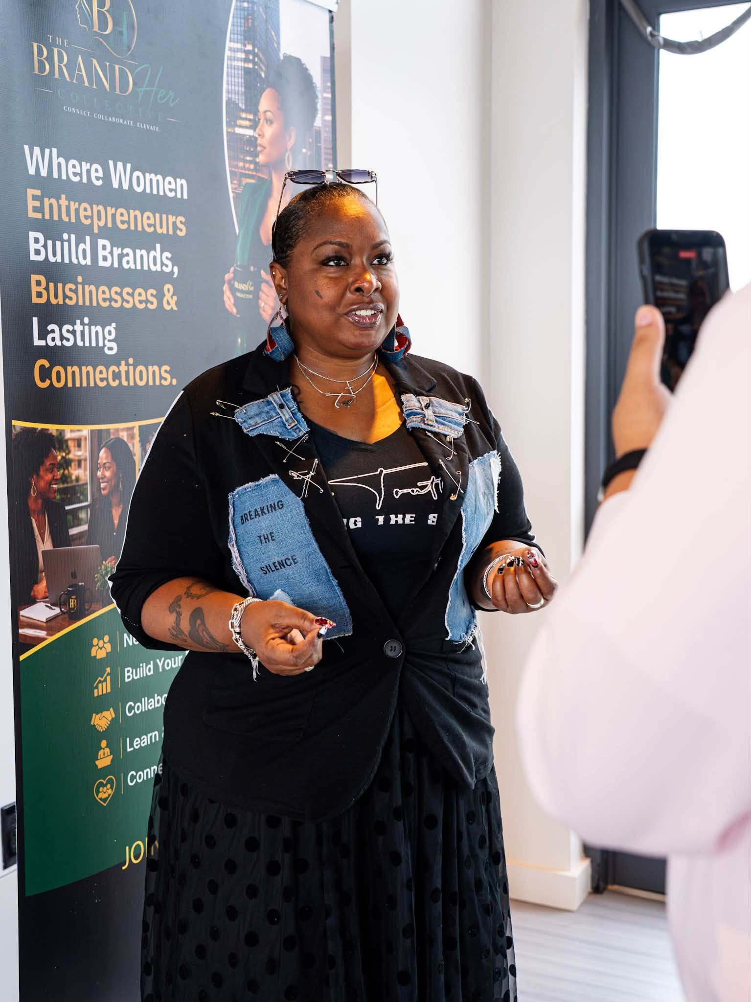Woman speaking in front of a banner, being photographed by another person, indoors with natural light.