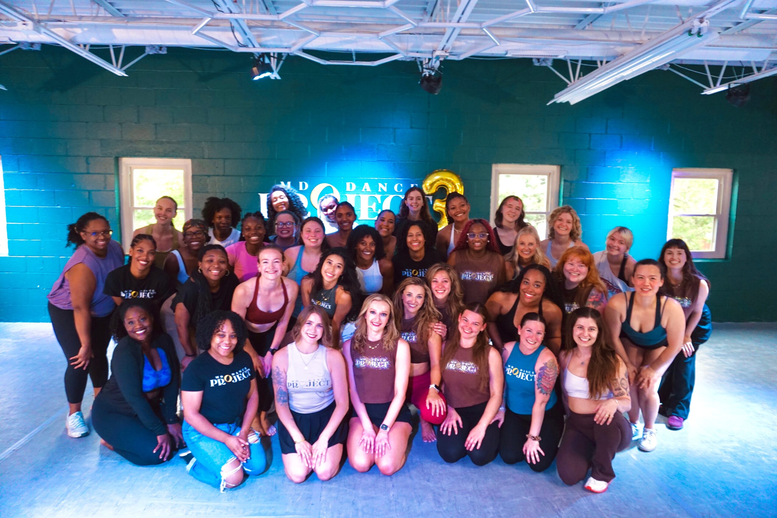 Group of young dancers posing together in a studio with blue lighting and framed pictures on the wall.