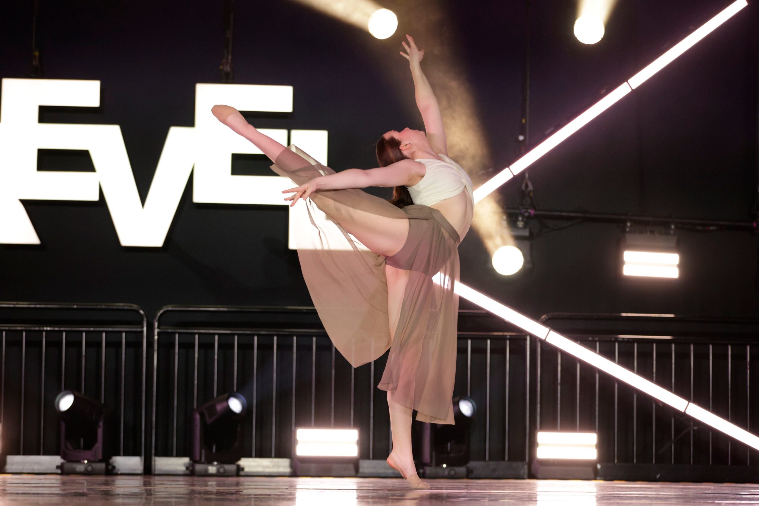 Ballet dancer performing on stage with lights and a black background, wearing a light-colored costume, in a graceful pose.