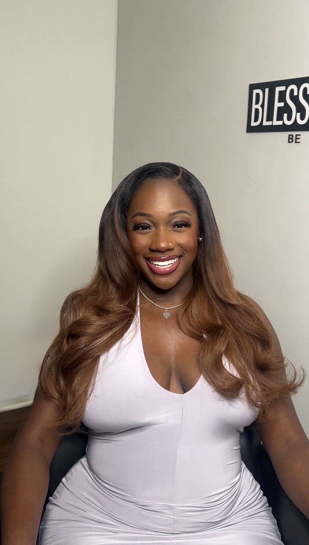 Smiling woman with long, wavy hair in a white top, sitting in front of a wall with a sign that says 'BLESS'.