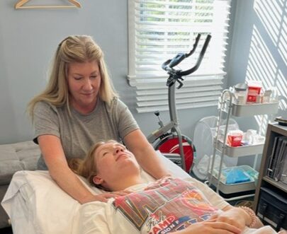 A woman with blonde hair cares for a person lying in bed in a room with sunlight through blinds.