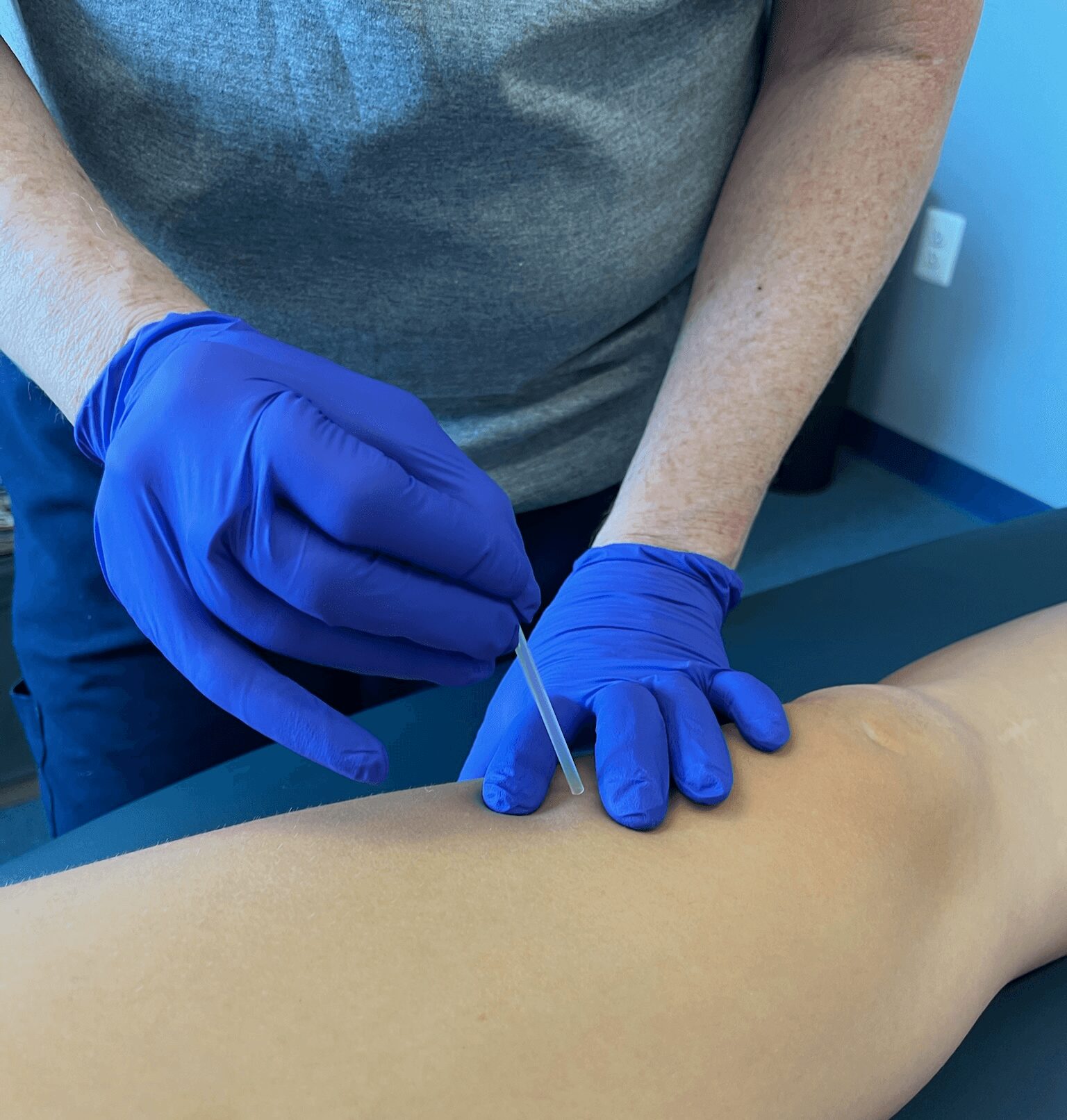 Person in blue gloves performs a medical procedure on a mannequin arm, inserting a needle or device.